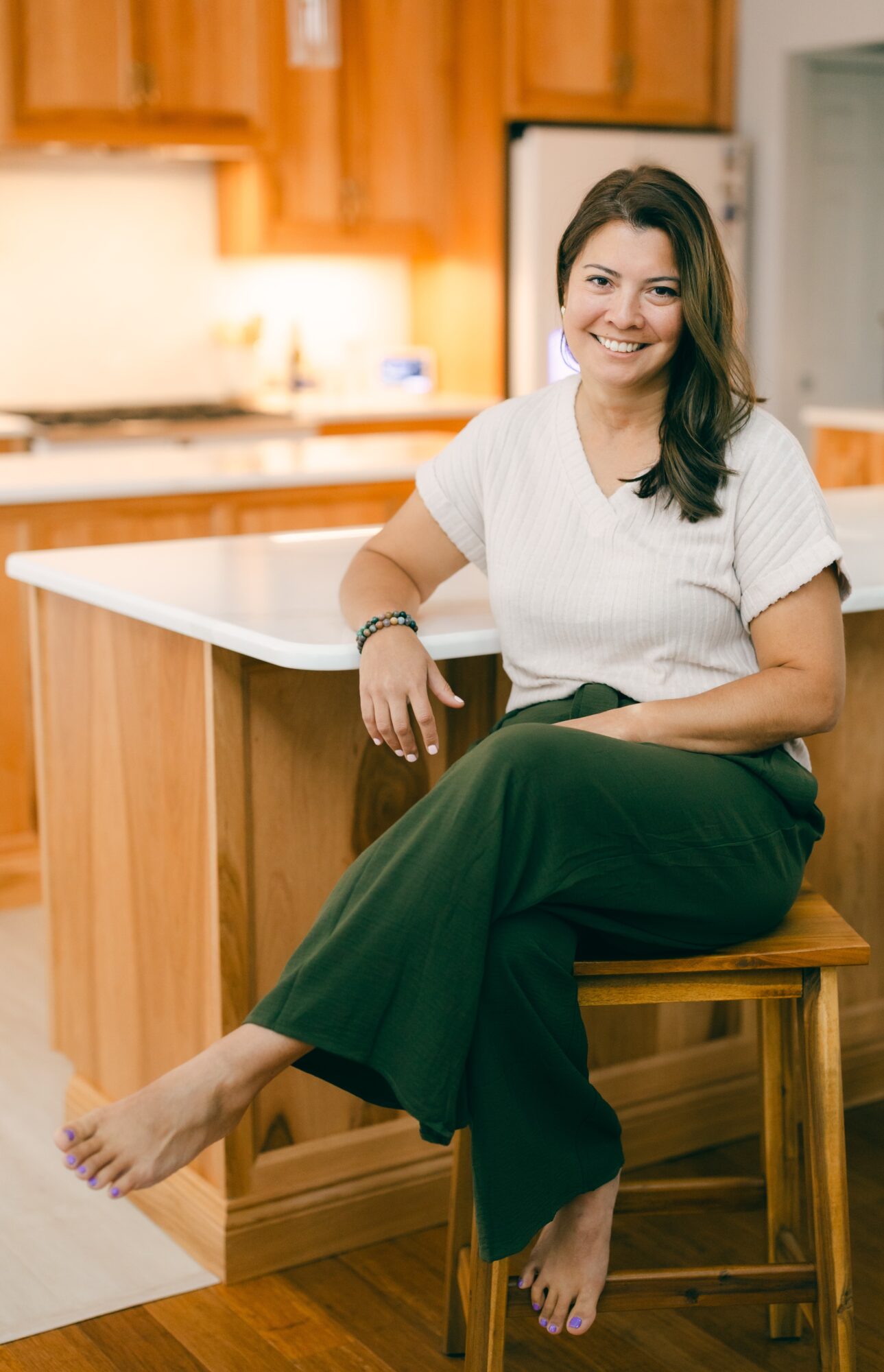 Woman sitting on a wooden chair in a kitchen, smiling, wearing a white top and dark pants, with a kitchen background.