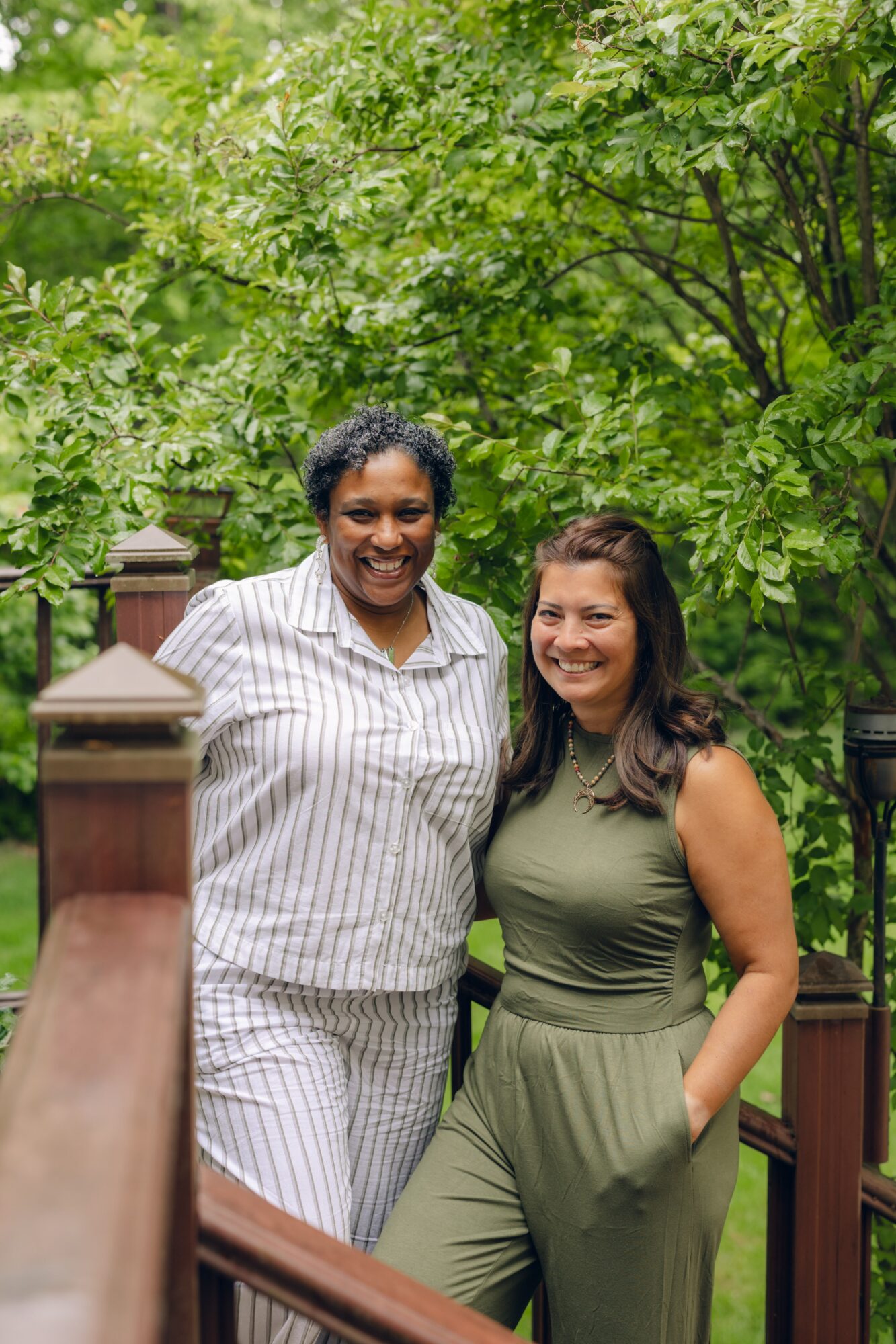 Two women stand outdoors in front of green trees, smiling, with a wooden fence and posts behind them.