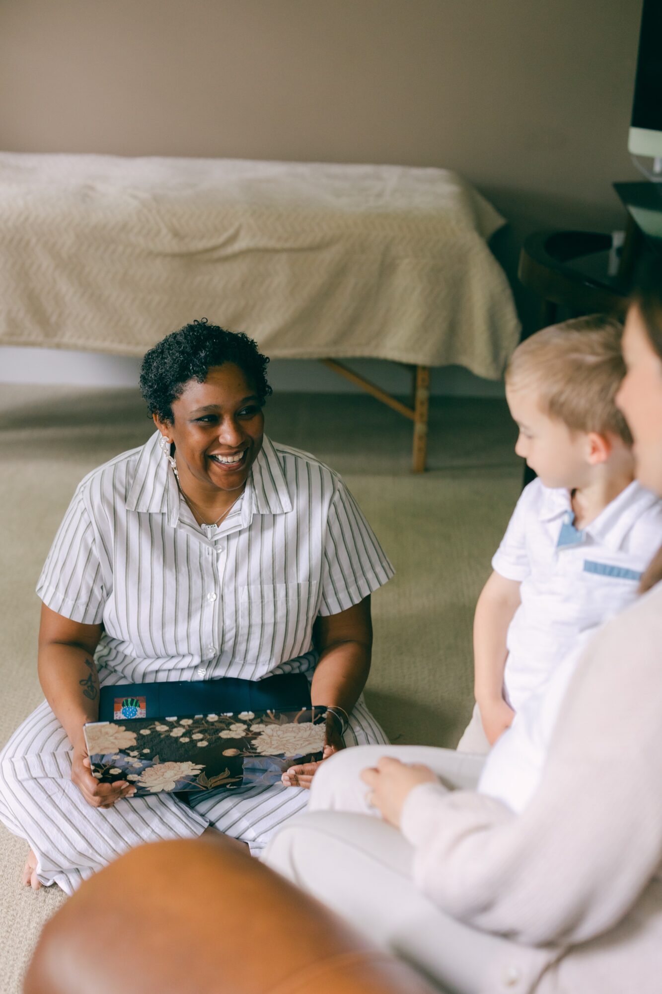 Woman smiling and holding a laptop, engaging with two children in a room with a bed in the background.