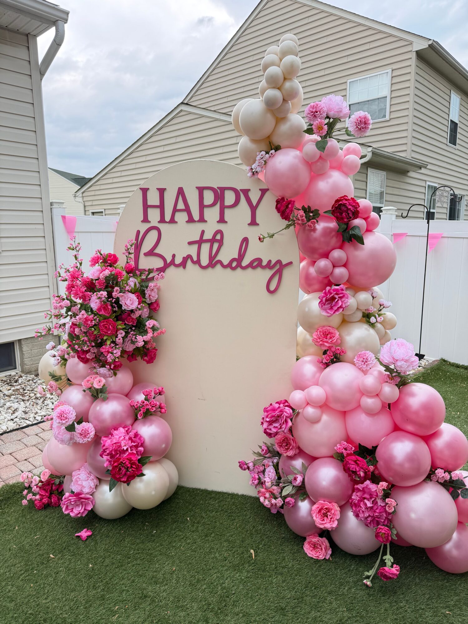 Birthday celebration with pink and white balloons and floral arrangements outside a house, with a 'Happy Birthday' sign.