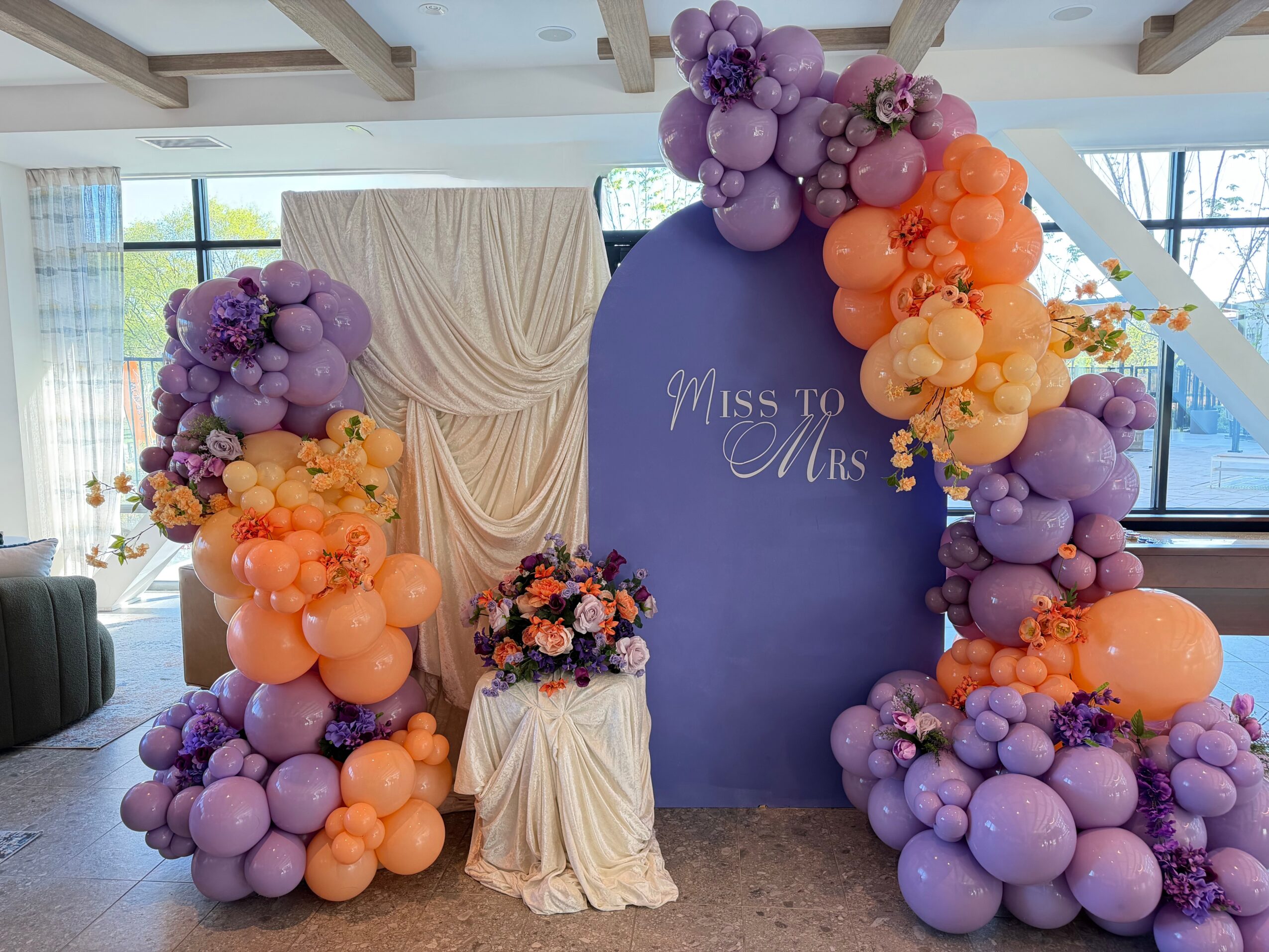 Decorative balloon arch with purple and orange balloons, a floral arrangement on a small table, and a backdrop with 'Miss to Mrs' text.