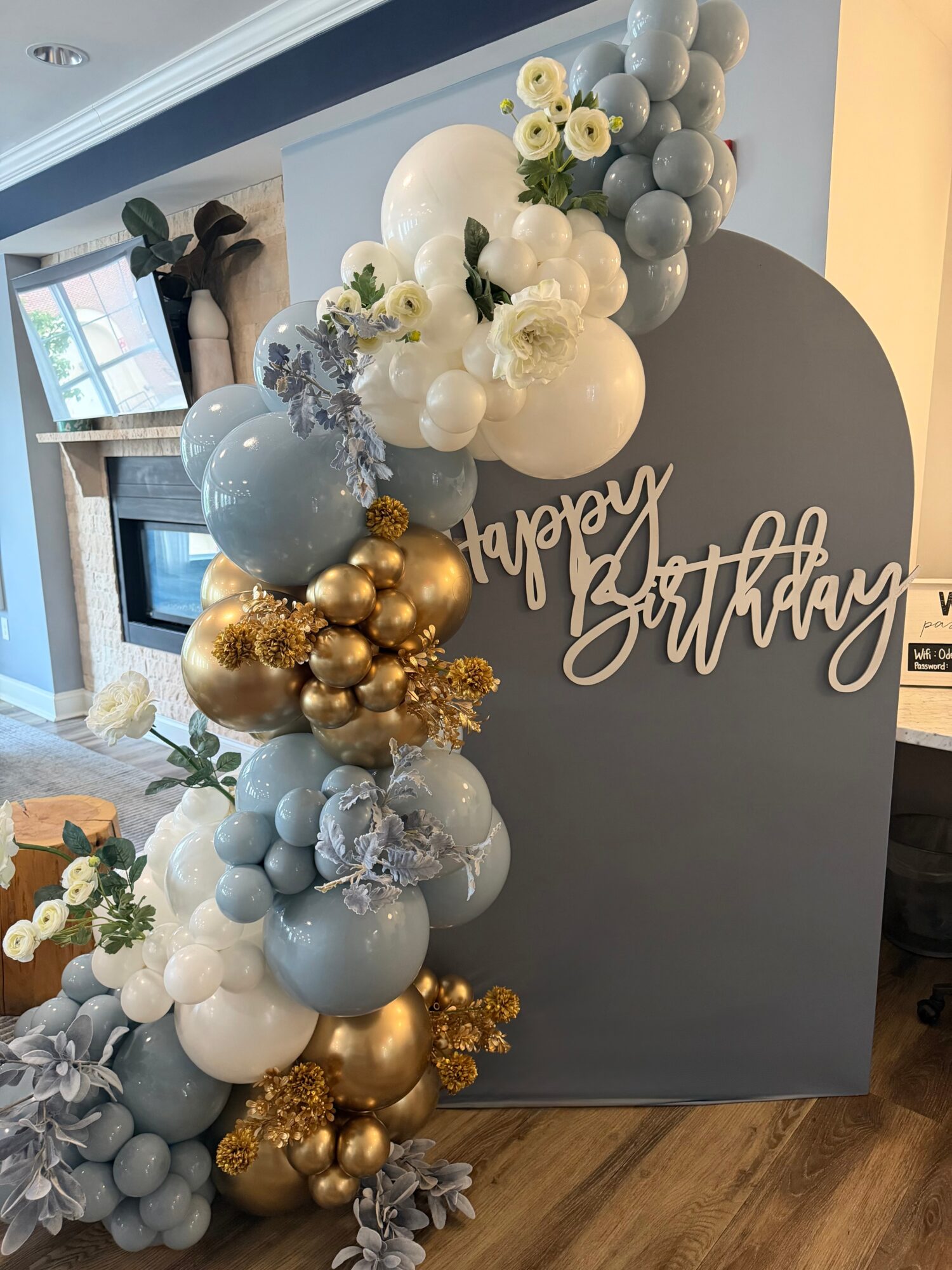 Decorative birthday display with balloons in white, blue, gray, and gold, and a gray backdrop with 'happy birthday' sign.