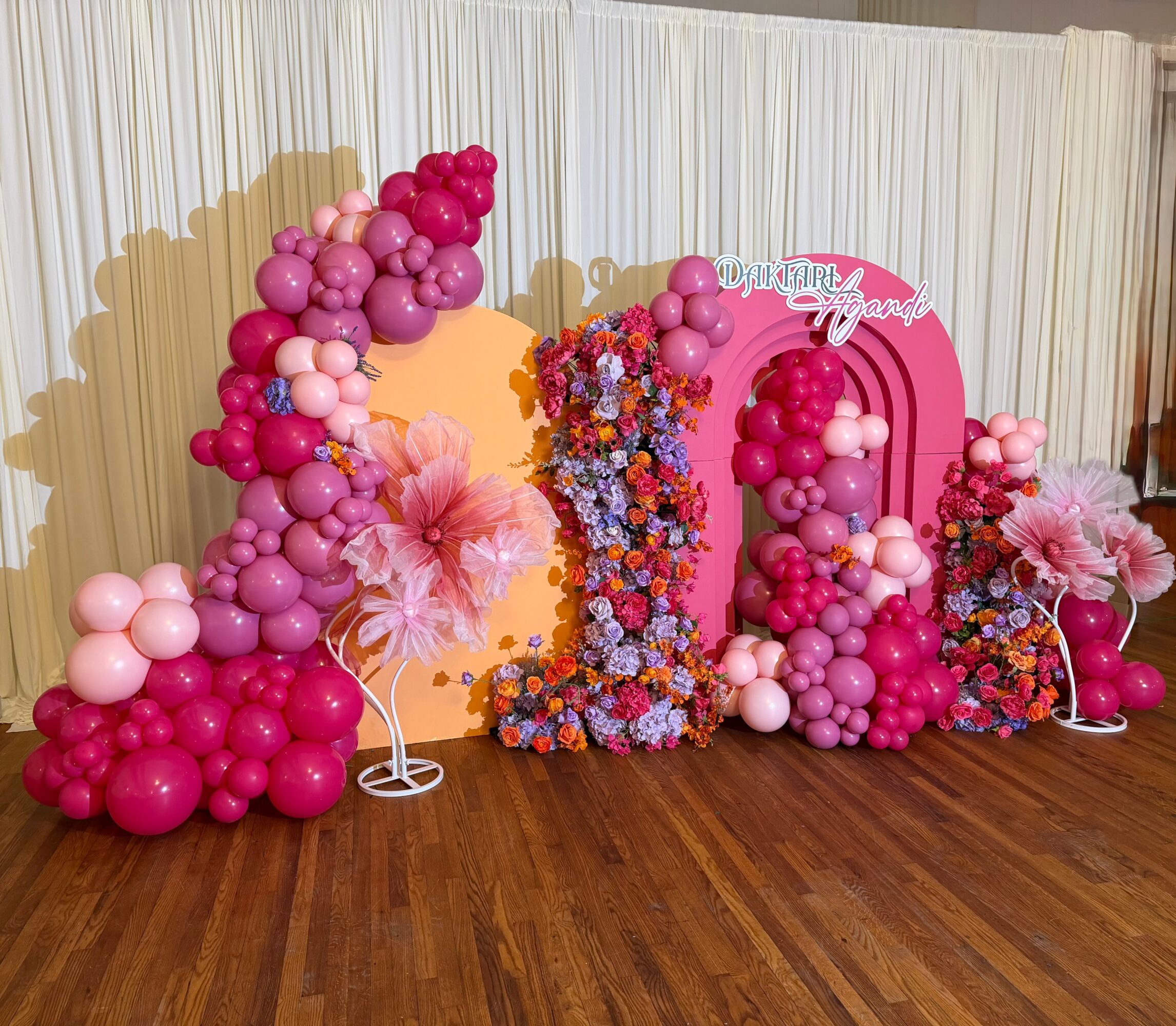 Decorative pink and purple balloon arrangement with flowers and a pink arch, on a wooden floor with white curtains behind.