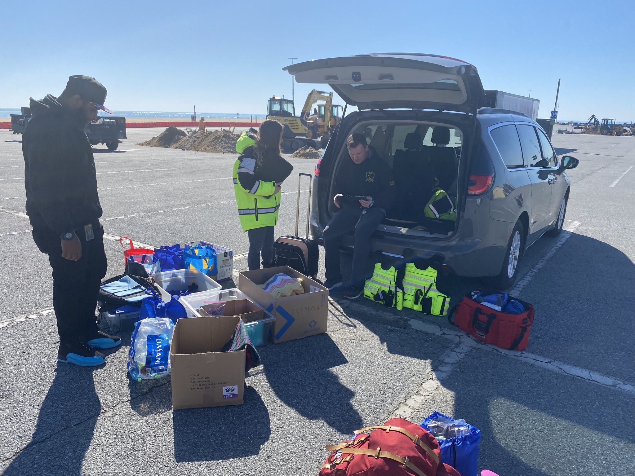 People gather around an open vehicle trunk with supplies and boxes on a parking lot, with a clear sky and distant boats.