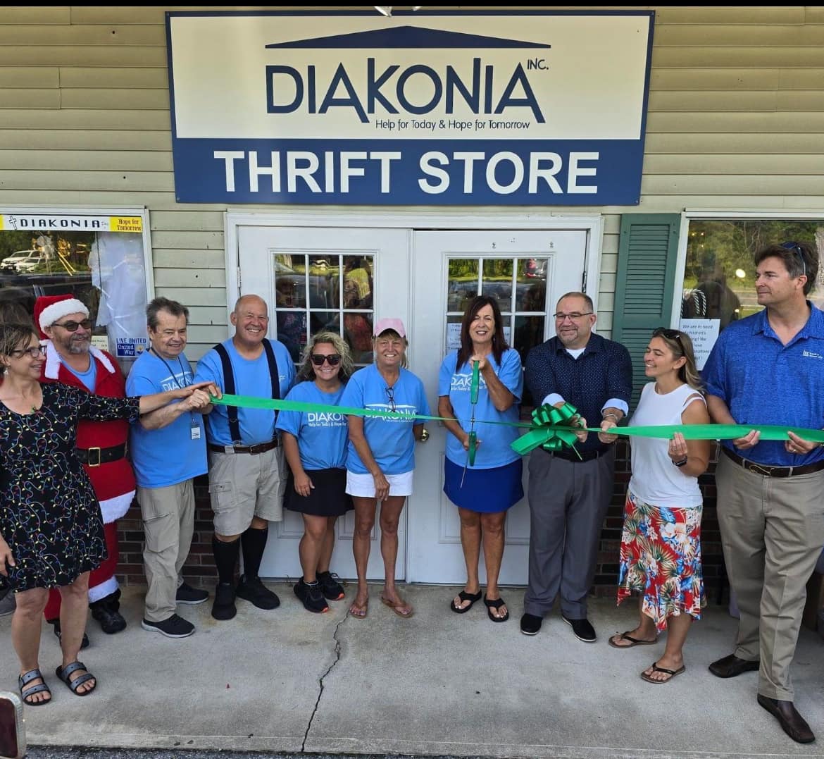Group of people at ribbon-cutting event outside thrift store, holding scissors and ribbon, smiling, in front of store entrance.