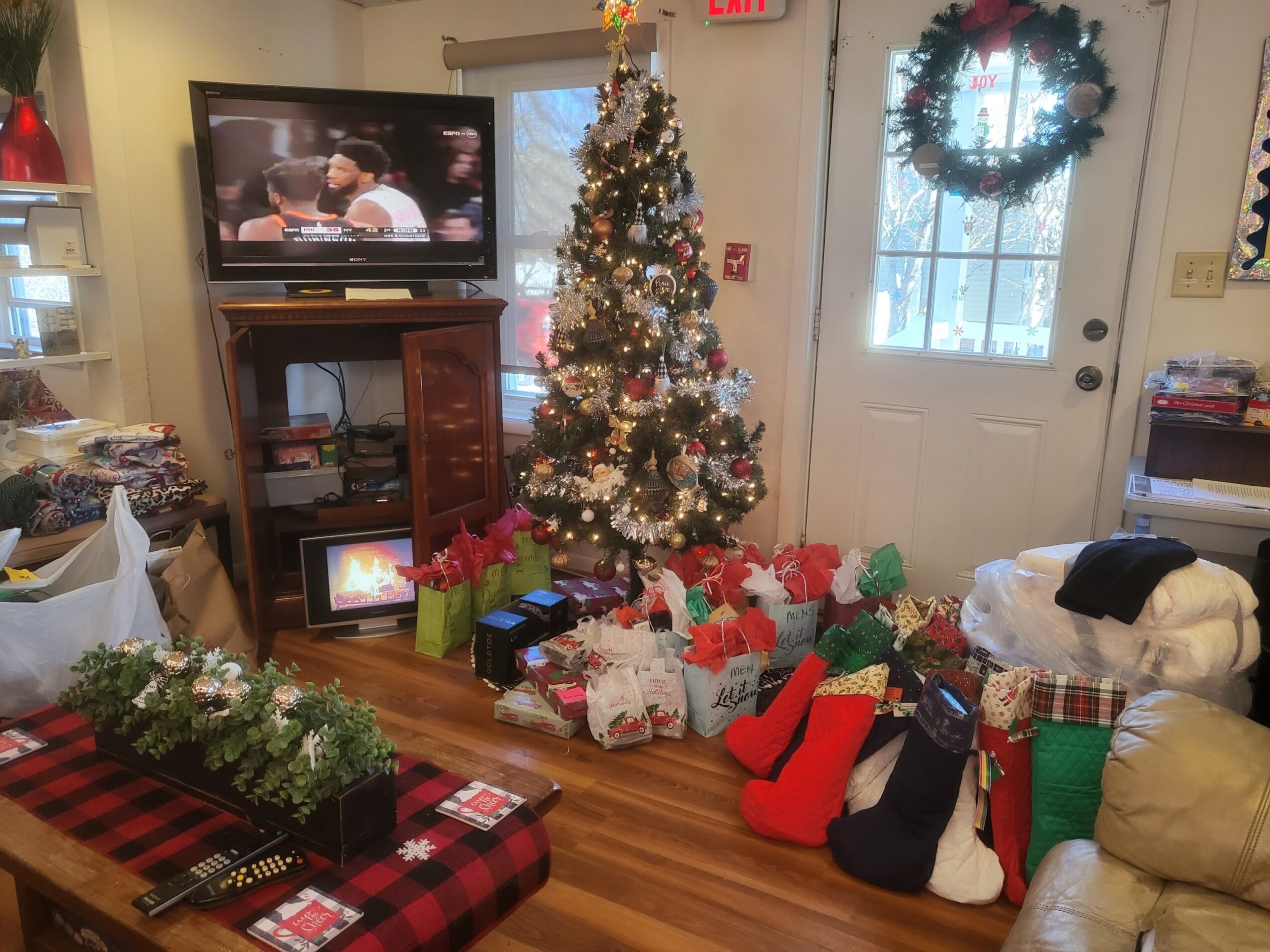 Christmas tree decorated with ornaments and lights, surrounded by wrapped presents and stockings, in a cozy room with a door and window.