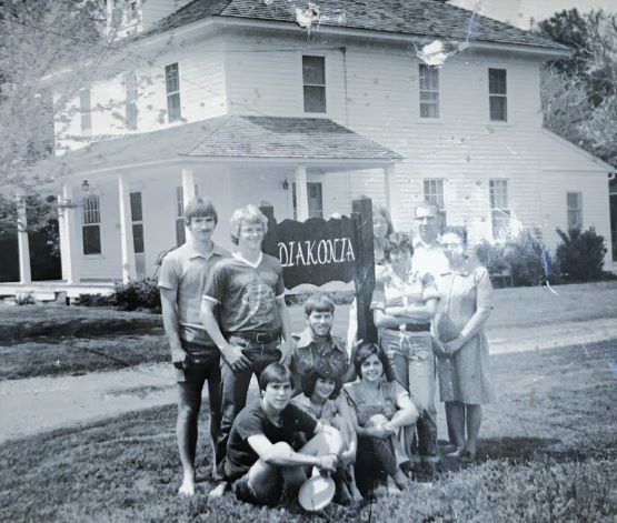 Group of seven people, including children and adults, standing and sitting in front of a house with a sign that reads 'DIAKONA'.