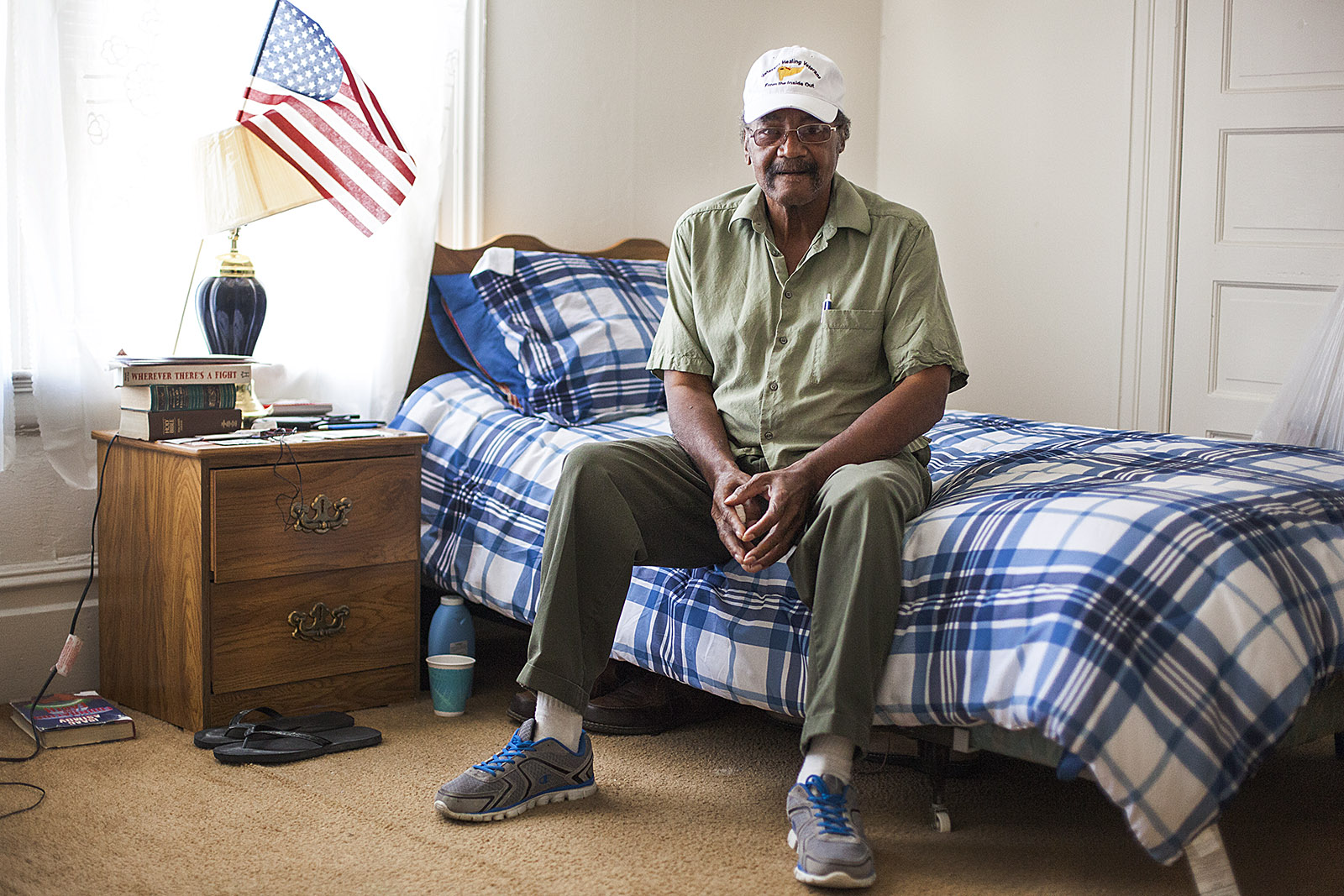 Older man sitting on a bed in a bedroom, smiling, with an American flag on a nightstand, plaid bedding, and a window with curtains.