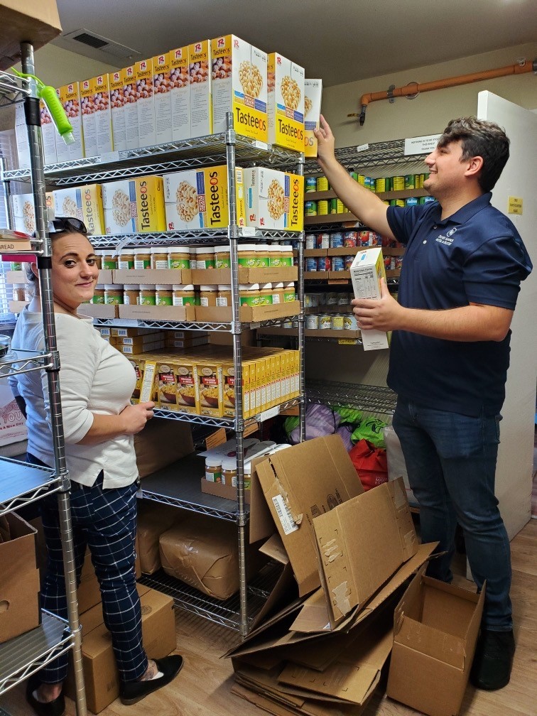 Two people in a storage room with shelves of cereal boxes, one reaching for a box, the other holding a box, surrounded by boxes and packaging materials.