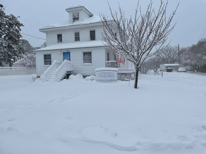 White house with stairs, surrounded by snow, with a leafless tree in front, and a sign near the tree.