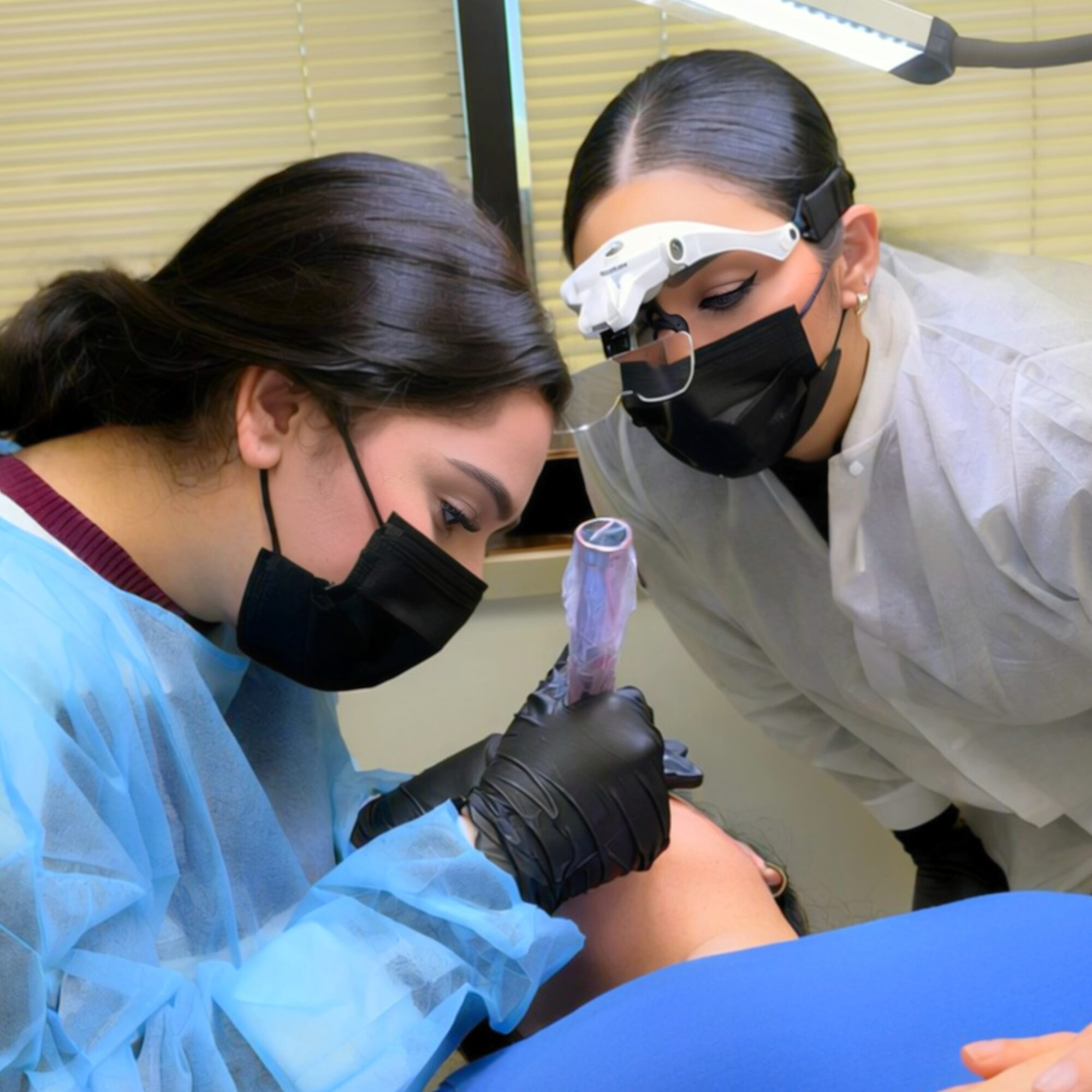 Two healthcare professionals, wearing masks and protective gear, perform a procedure on a patient in a clinical setting.