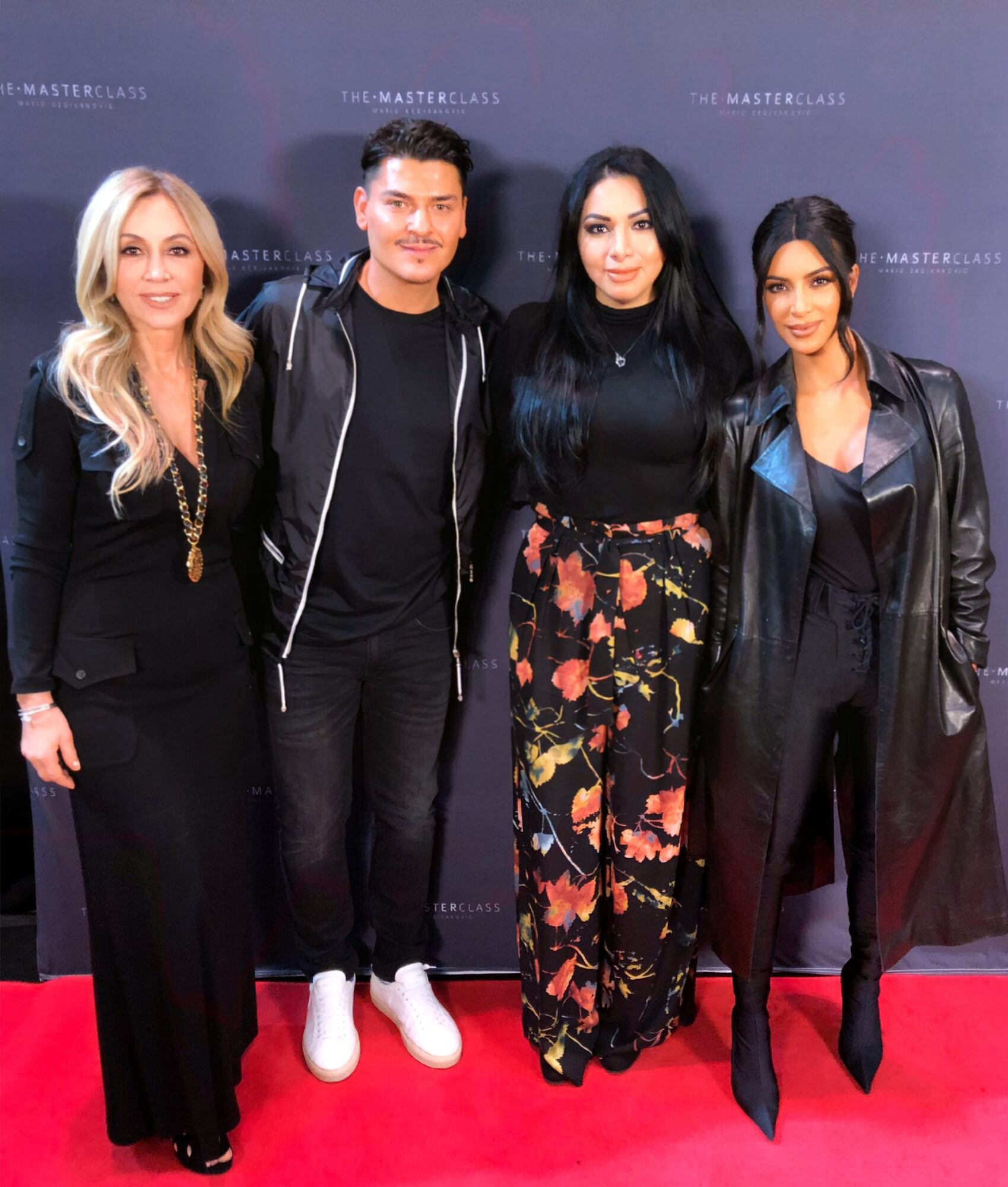 Four people standing on a red carpet in front of a dark backdrop, posing for a photo, dressed in black and floral clothing.