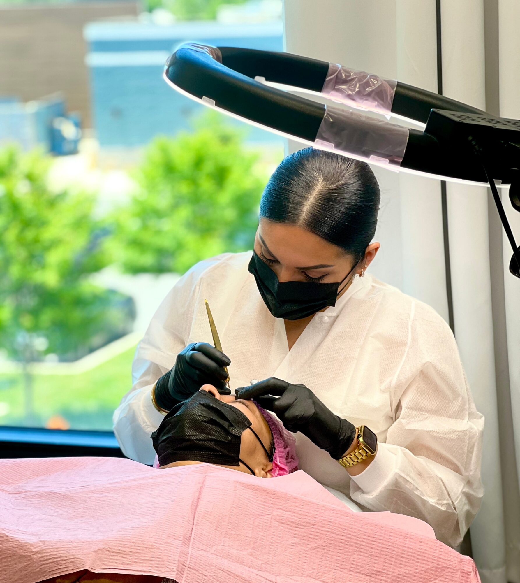 A person wearing a black mask, gloves, and white coat performs a procedure on a patient with a pink cloth, near a window with greenery outside.