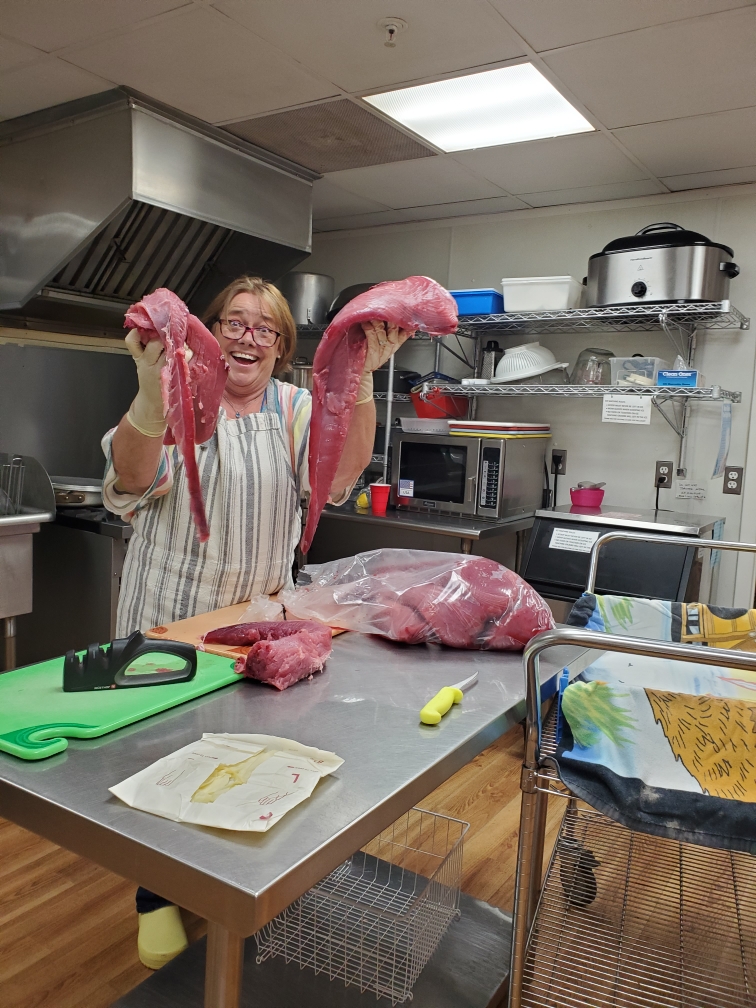 Person in a kitchen holding two large pieces of raw meat, smiling, with kitchen appliances and shelves in the background.