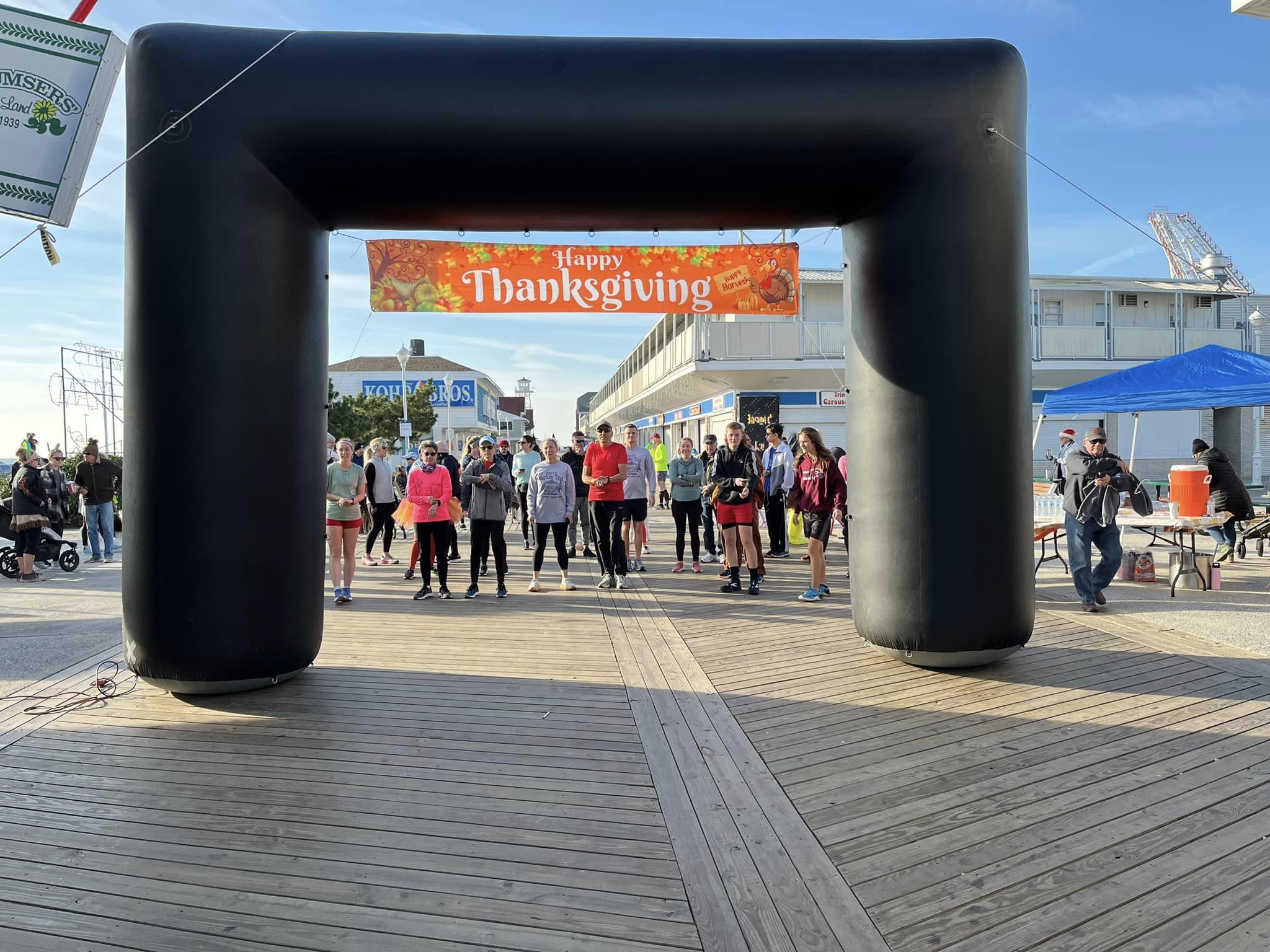 People gather under a large black inflatable arch with a Thanksgiving banner, on a wooden boardwalk outdoors.