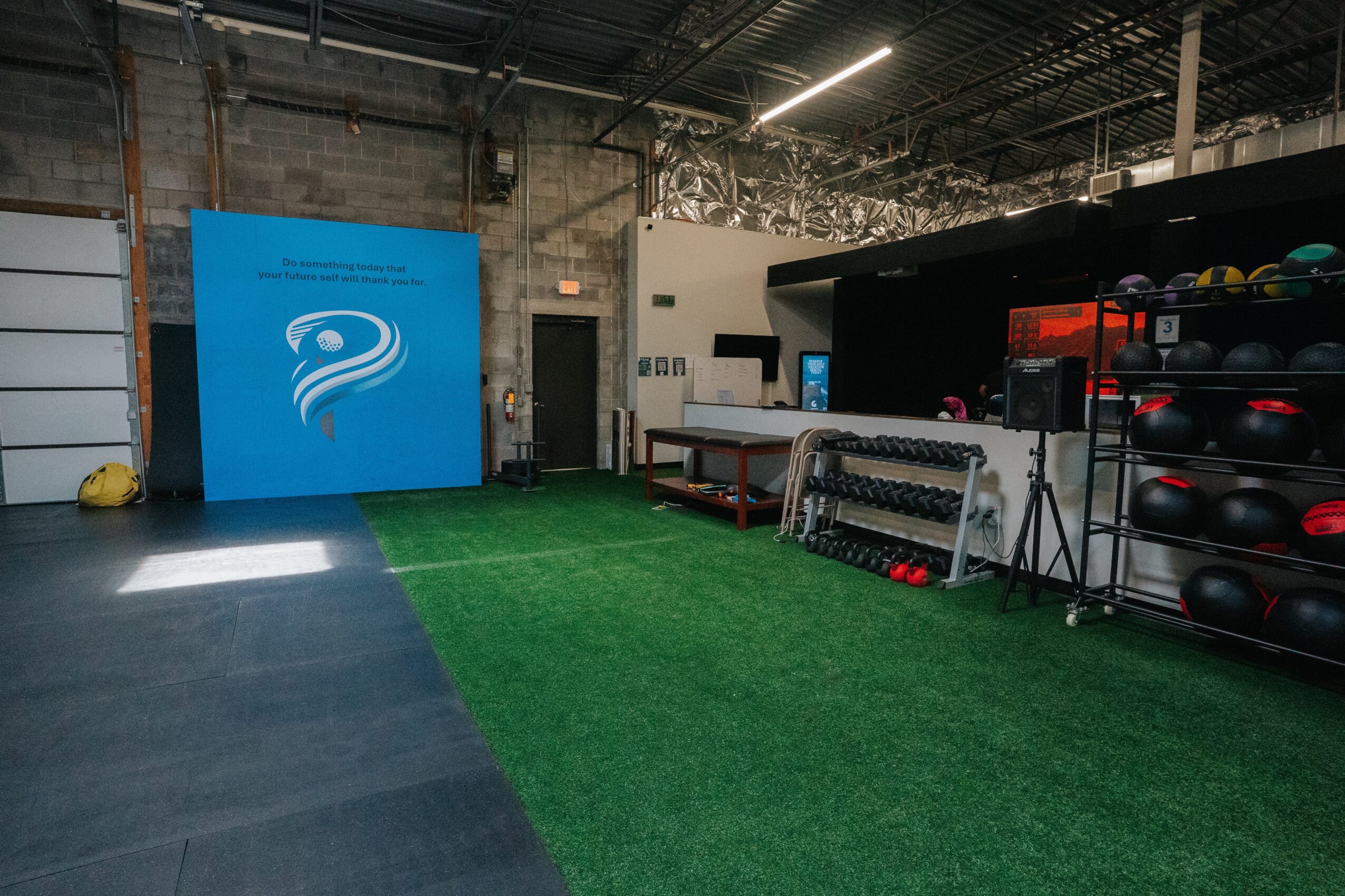 Indoor gym area with artificial grass, fitness equipment, and a blue wall with a logo, in a warehouse-style space.