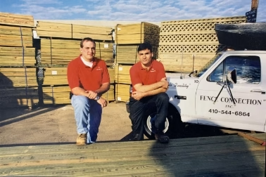 Two men kneeling outdoors near stacked wooden pallets and a white vehicle with text, in bright sunlight.