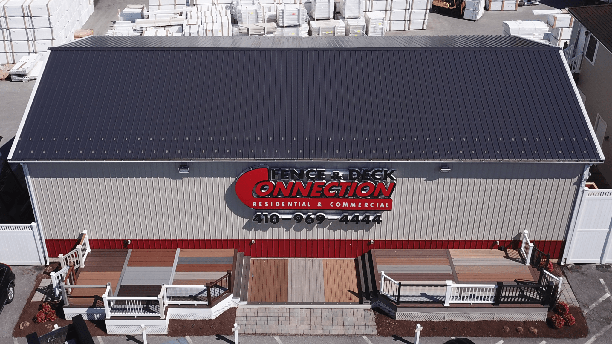 Building with a sign reading 'FENCE & DECK CONNECTION' and a black roof, with outdoor seating area in front.