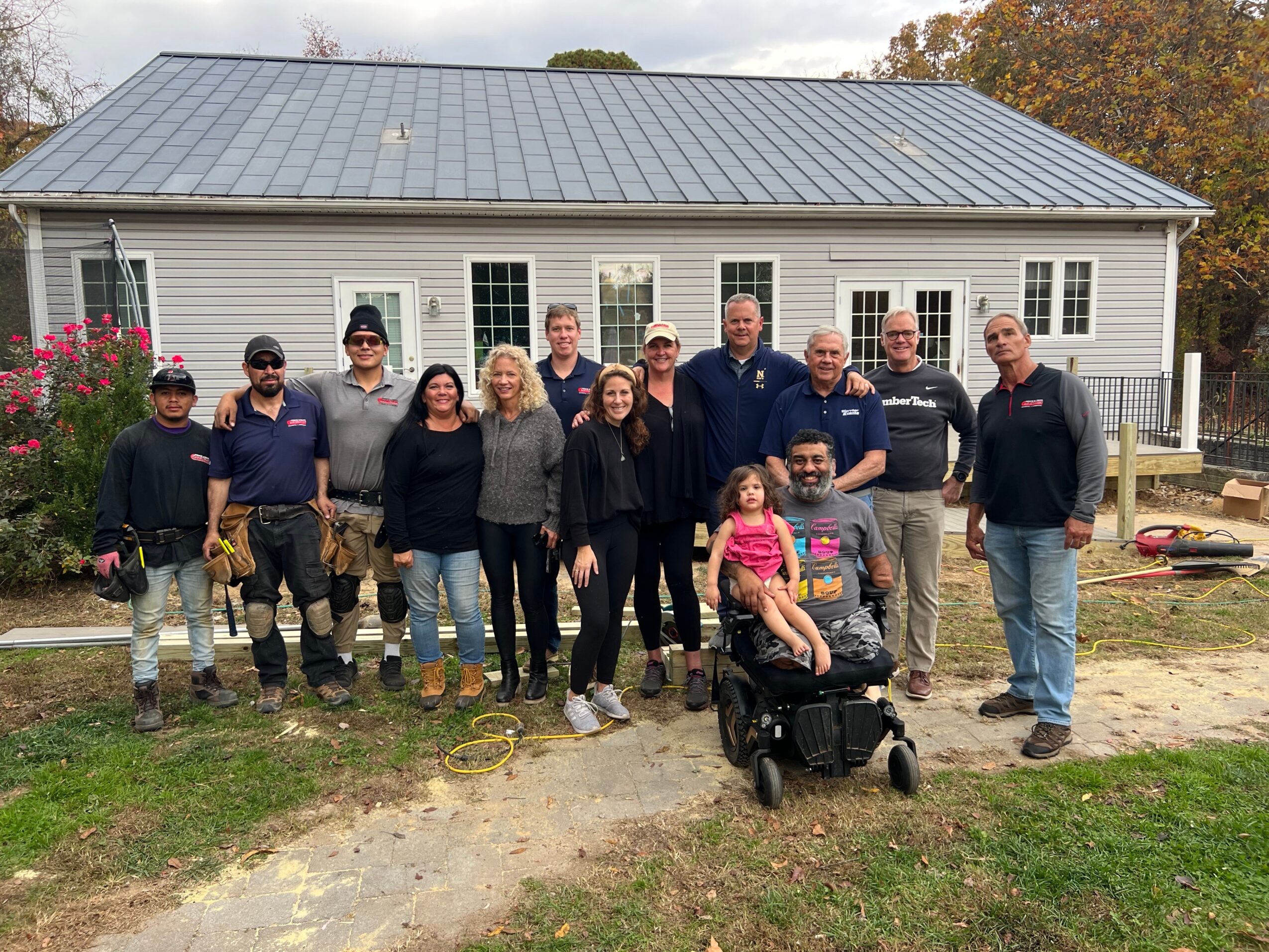 Group of 14 people, including children, standing outside a house with a metal roof, some smiling, in a yard with trees in the background.