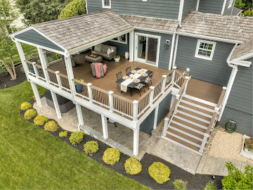 Elevated outdoor deck with dining table, chairs, and seating area, attached to a blue house with stairs leading down.