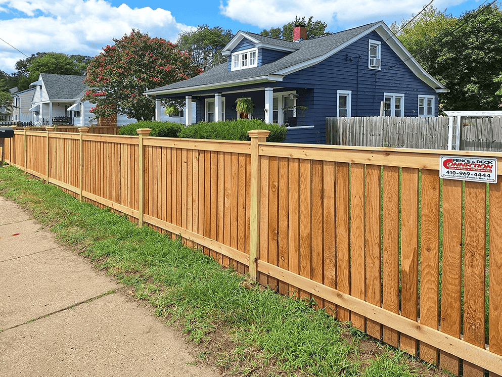 New wooden fence along sidewalk with a blue house and trees in the background.