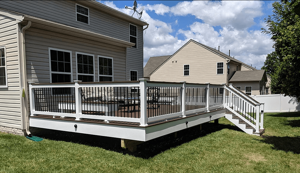 A raised outdoor deck with white railing and stairs, attached to a house, on a grassy yard under a partly cloudy sky.