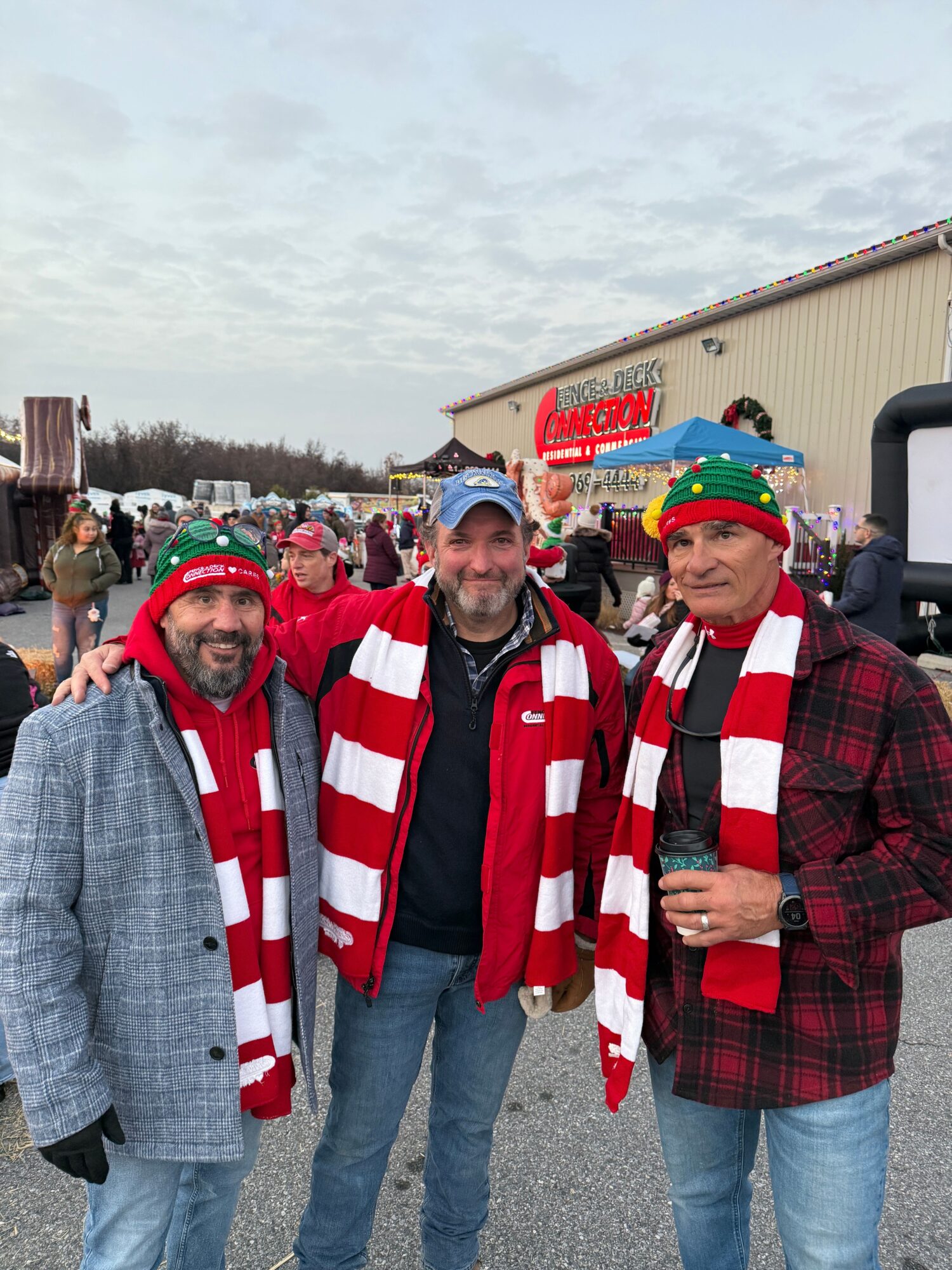 Three people dressed in Christmas-themed clothing standing outdoors during a holiday event, with a crowd and festive decorations in the background.