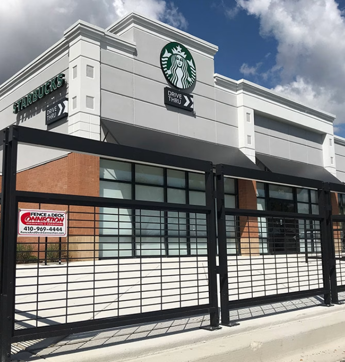 Starbucks store with logo, large windows, and a black metal fence in front, under a partly cloudy sky.
