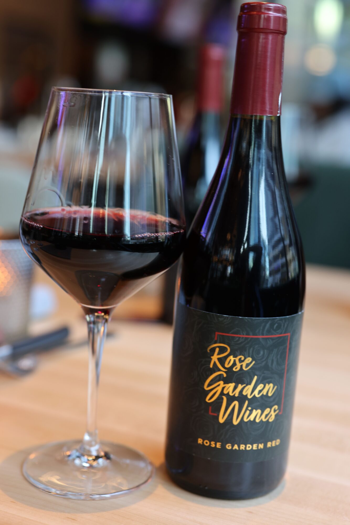 A glass of red wine on a wooden surface with a wine bottle nearby, background blurred.