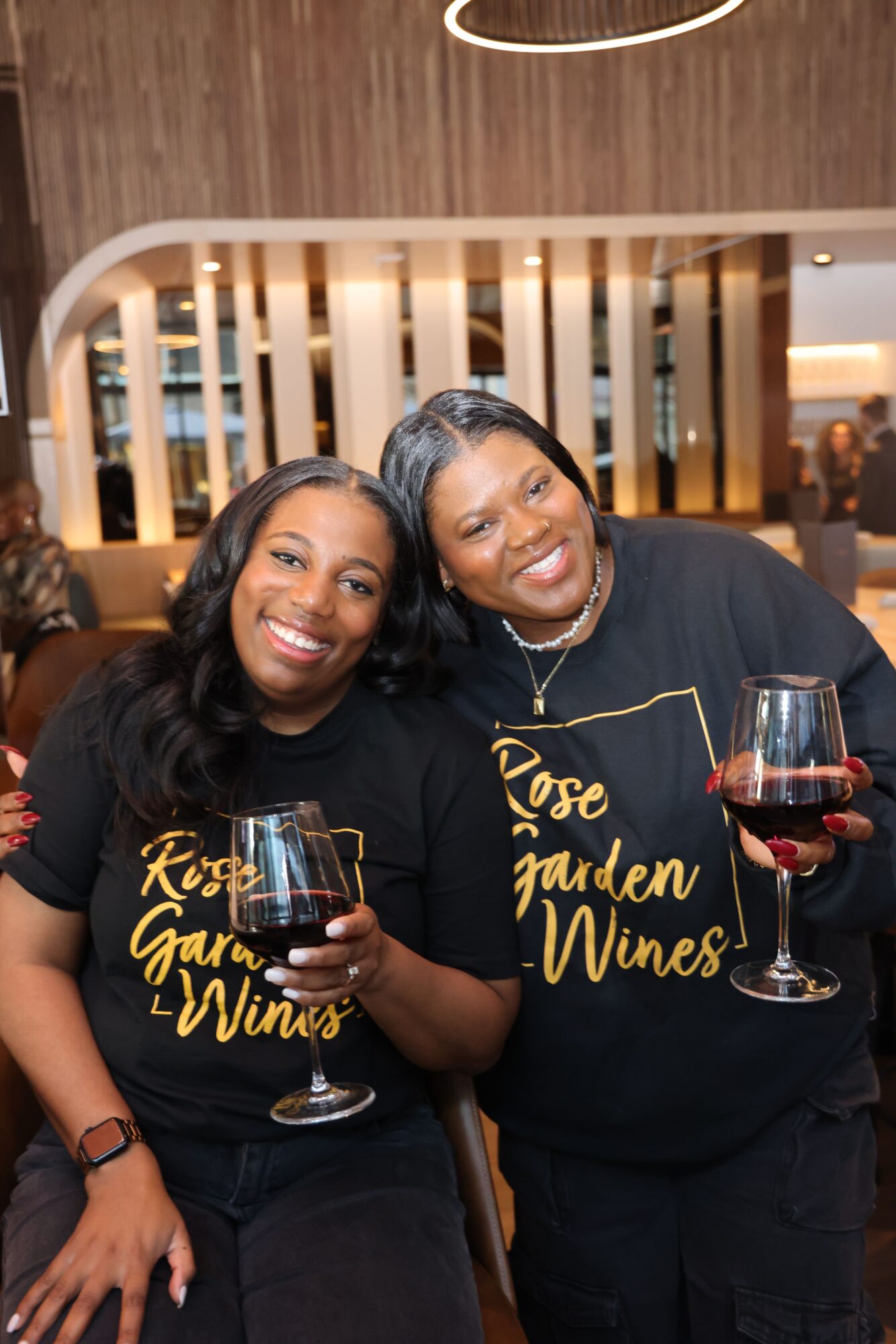 Two women smiling, holding wine glasses, sitting close together indoors with wooden decor in the background.