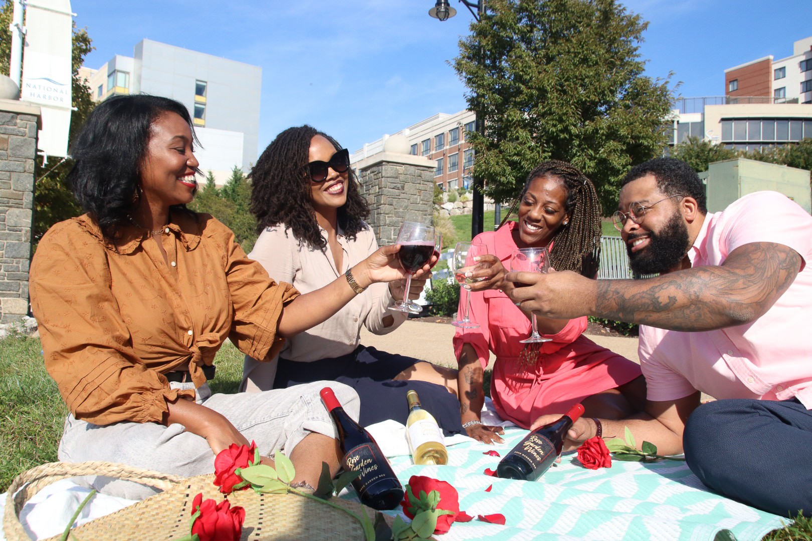 Four people sitting on a blanket outdoors, smiling, holding drinks, with bottles and roses, enjoying a sunny day.