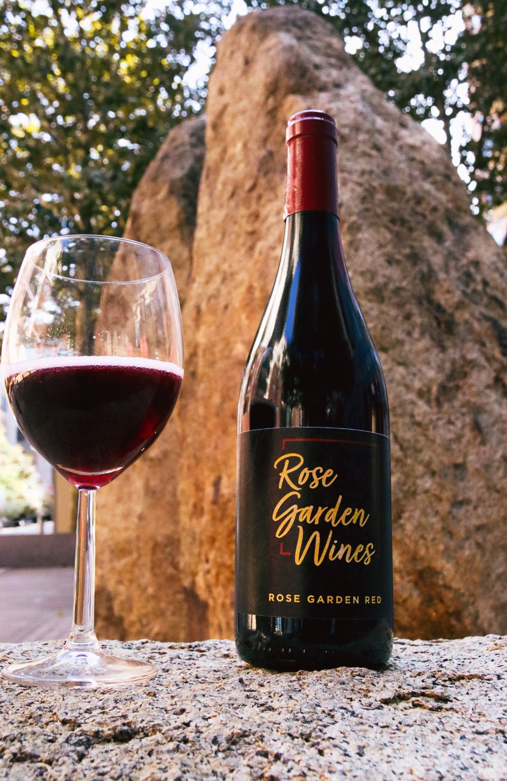 Bottle of Rose Garden Wines red wine and a glass of red wine outdoors with a large rock and trees in background