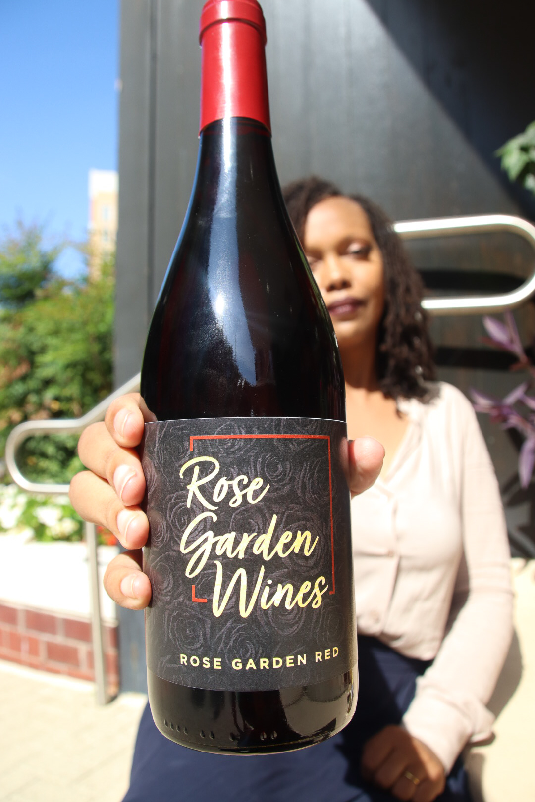 Person holding a bottle of Rose Garden Wines wine outdoors, with a woman and greenery in the background.