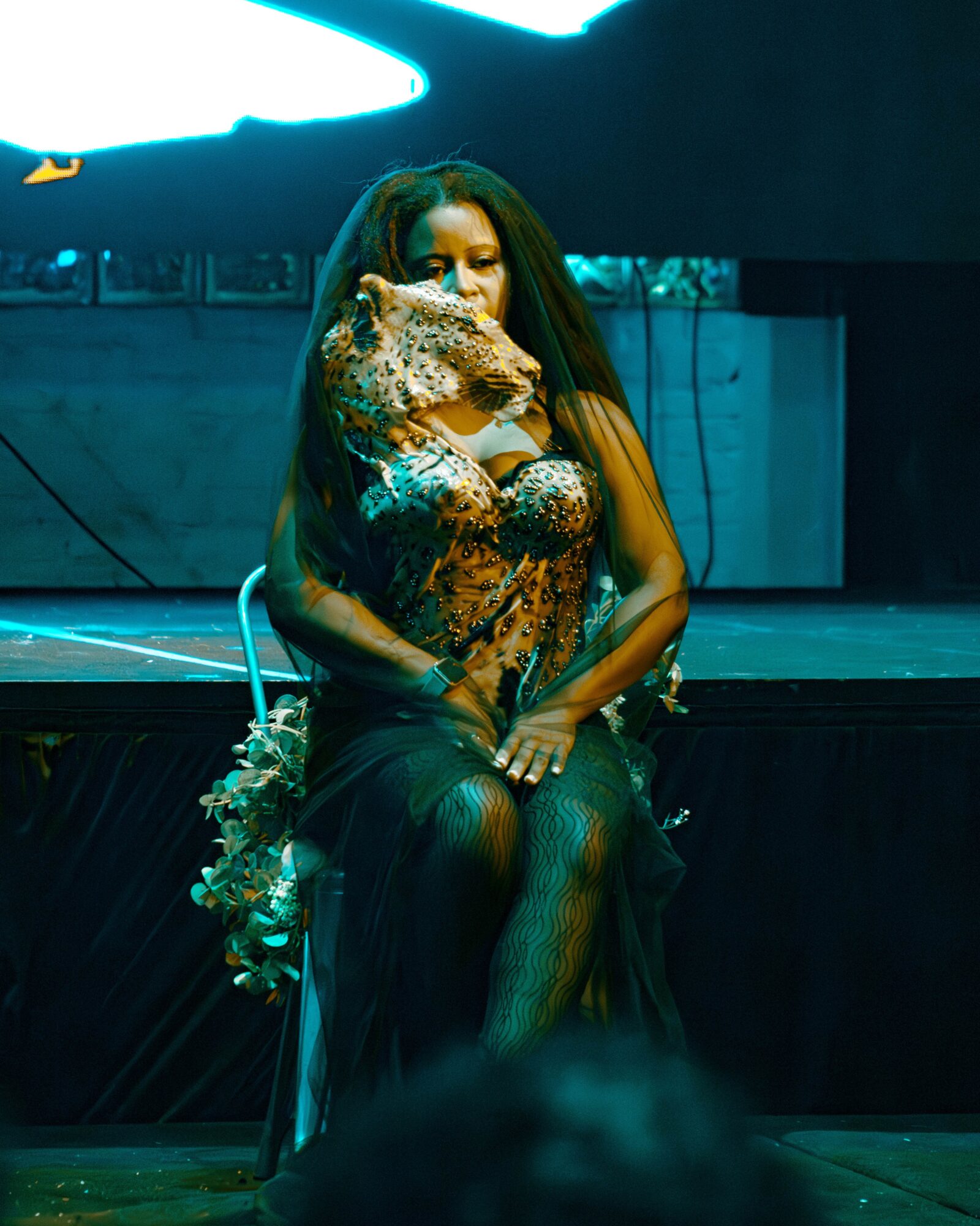 Woman sitting on stage, wearing a black dress, with a leopard-print face mask, and long dark hair, in a dark setting.
