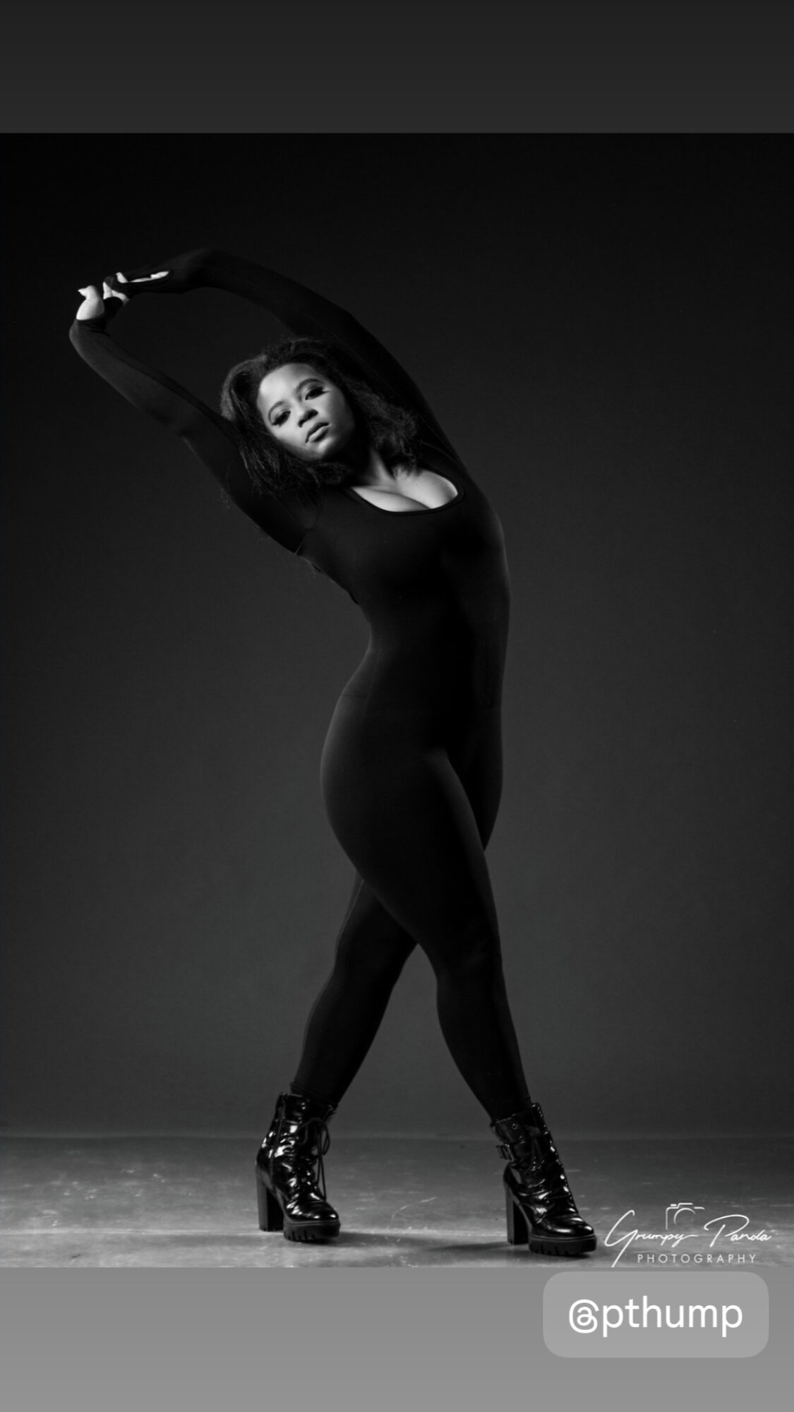 Woman in black outfit and boots poses with arms raised above head, standing on a dark stage.