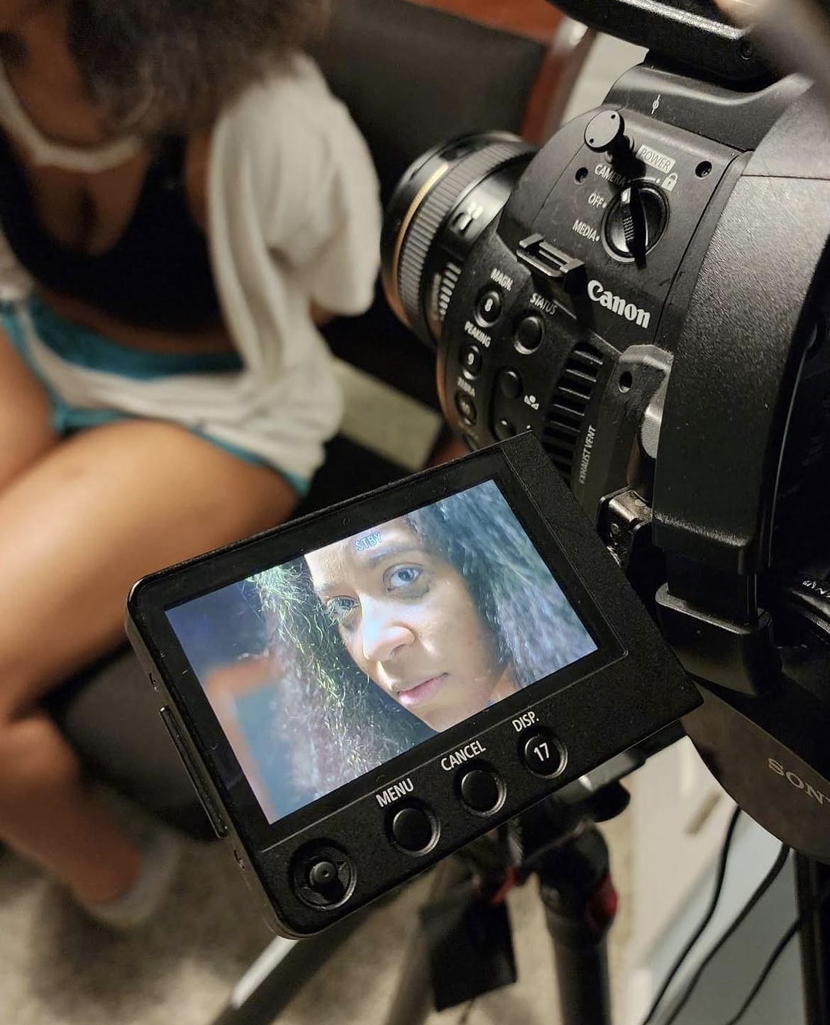 Close-up of a camera's LCD screen displaying a woman's face with curly hair, with a person blurred in the background.