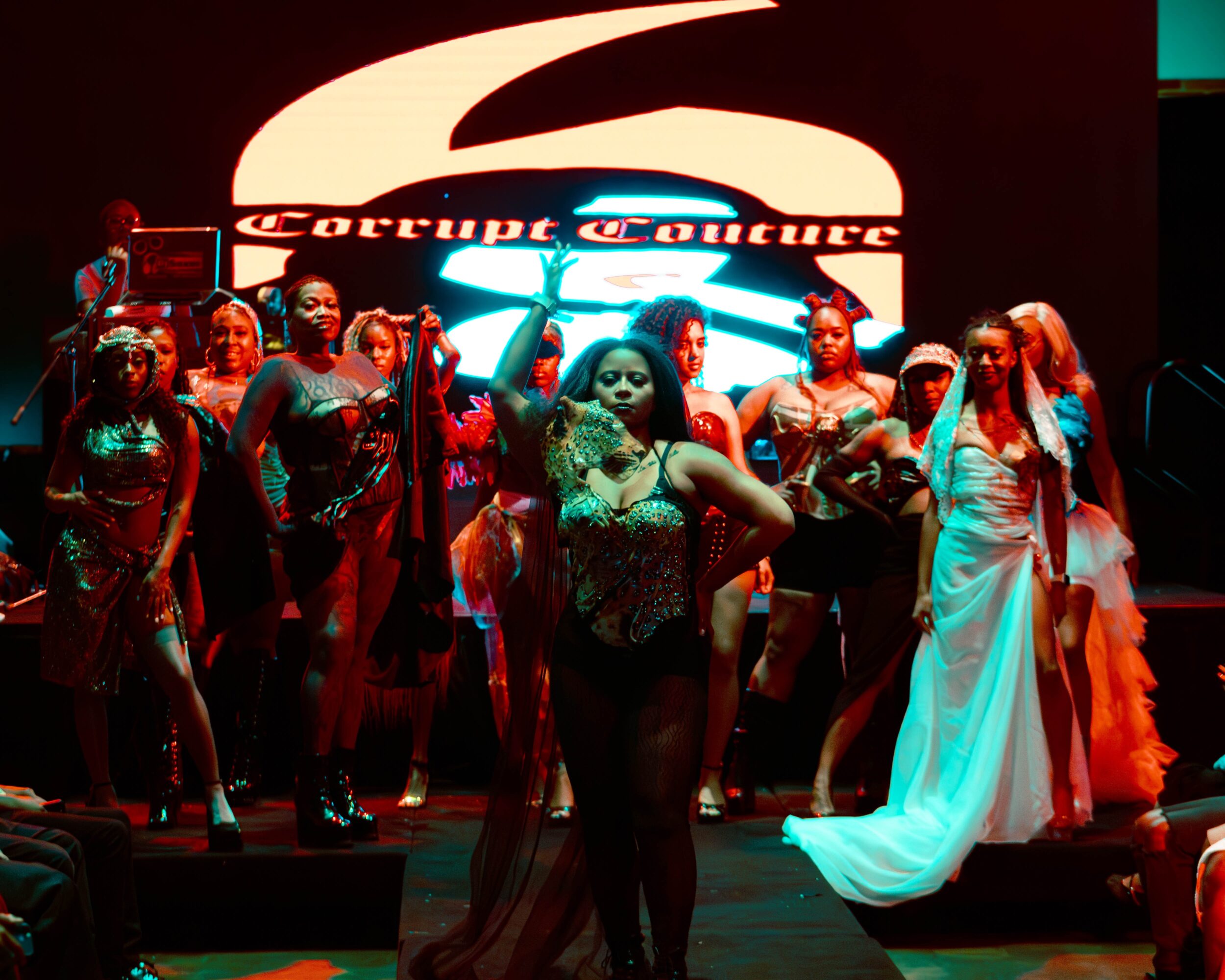 Group of women on stage with a large illuminated sign behind them, some wearing glamorous outfits, in a performance setting.