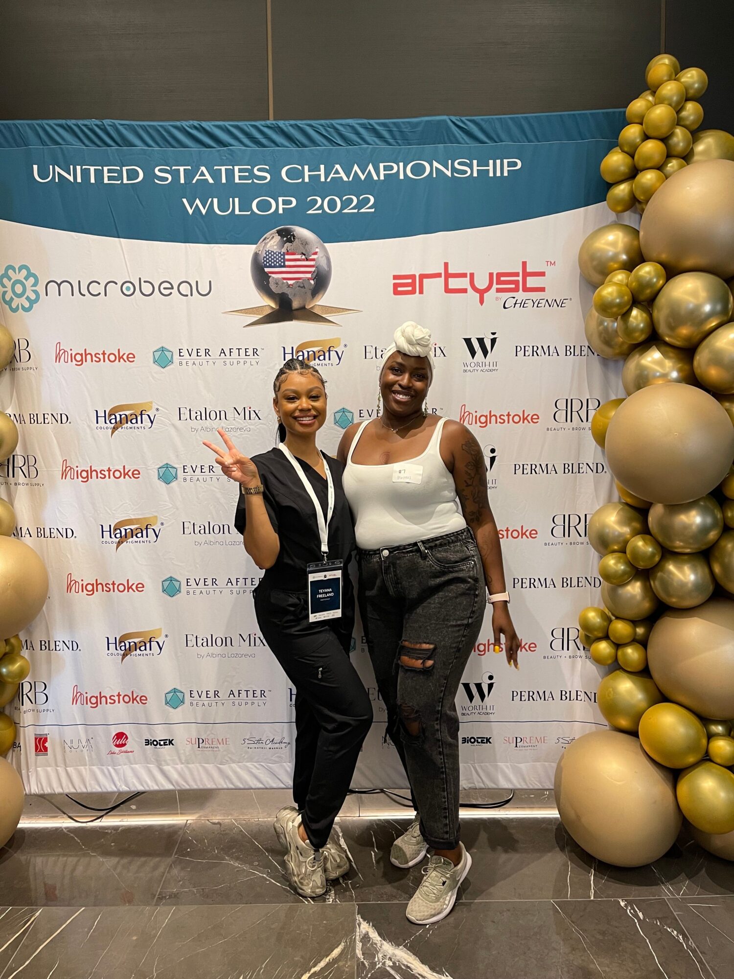 Two women standing in front of a decorated backdrop with gold and silver balloons, smiling at the camera.