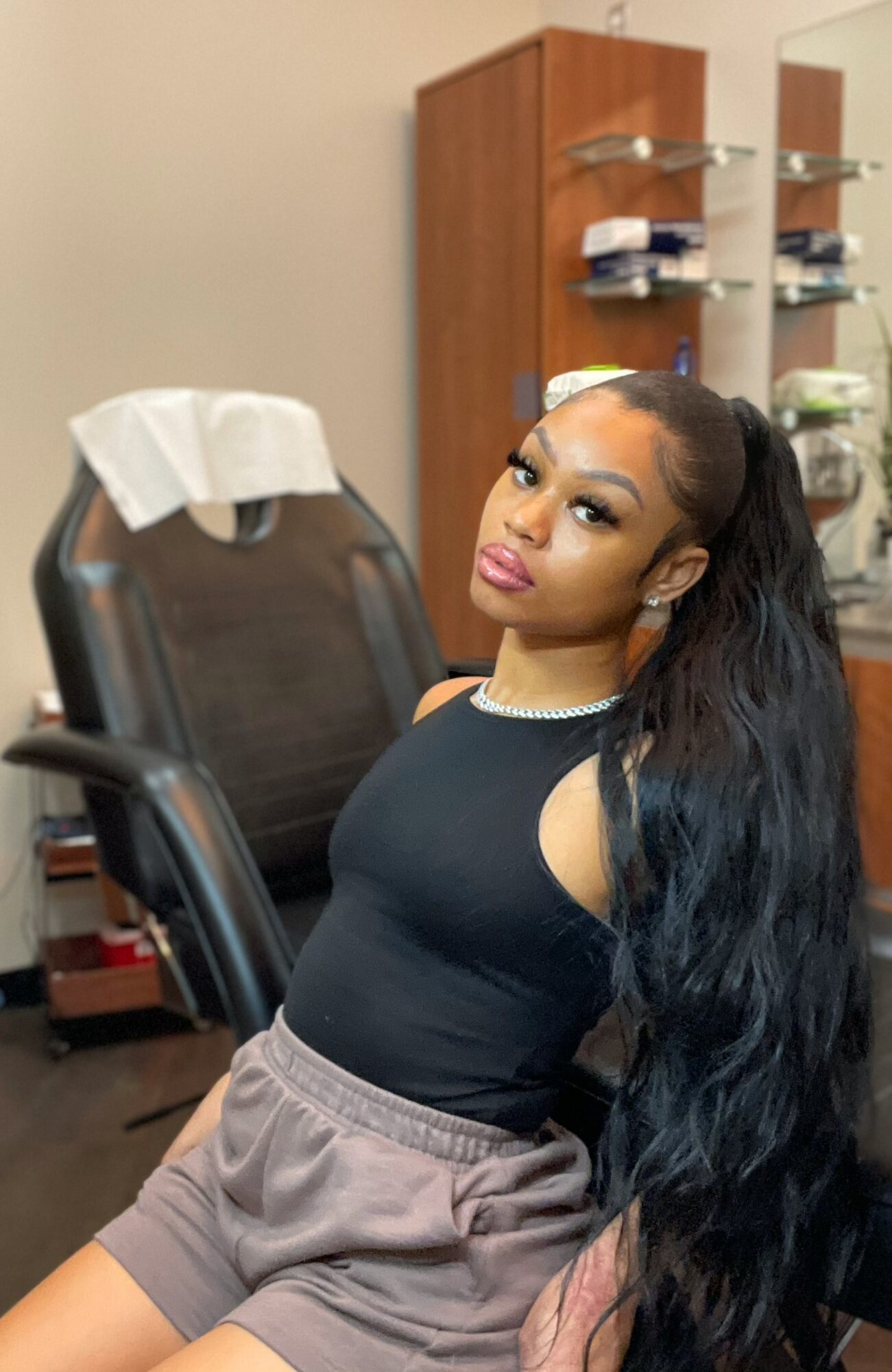 Young woman with long black hair sitting in a medical chair, looking at the camera, in a clinic room.