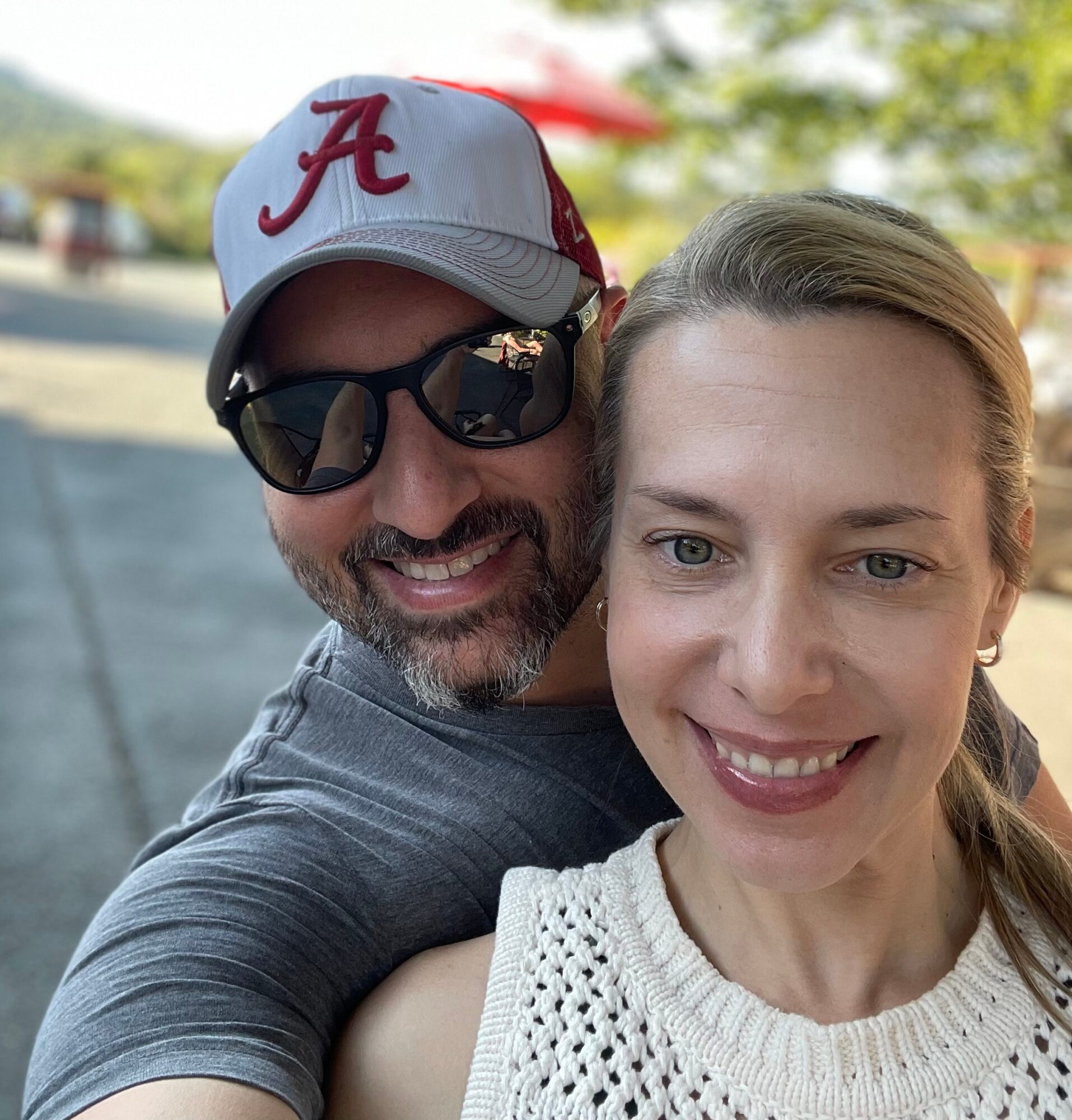 A man and woman smiling outdoors, man wearing sunglasses and a baseball cap, woman with blonde hair and a white sweater.