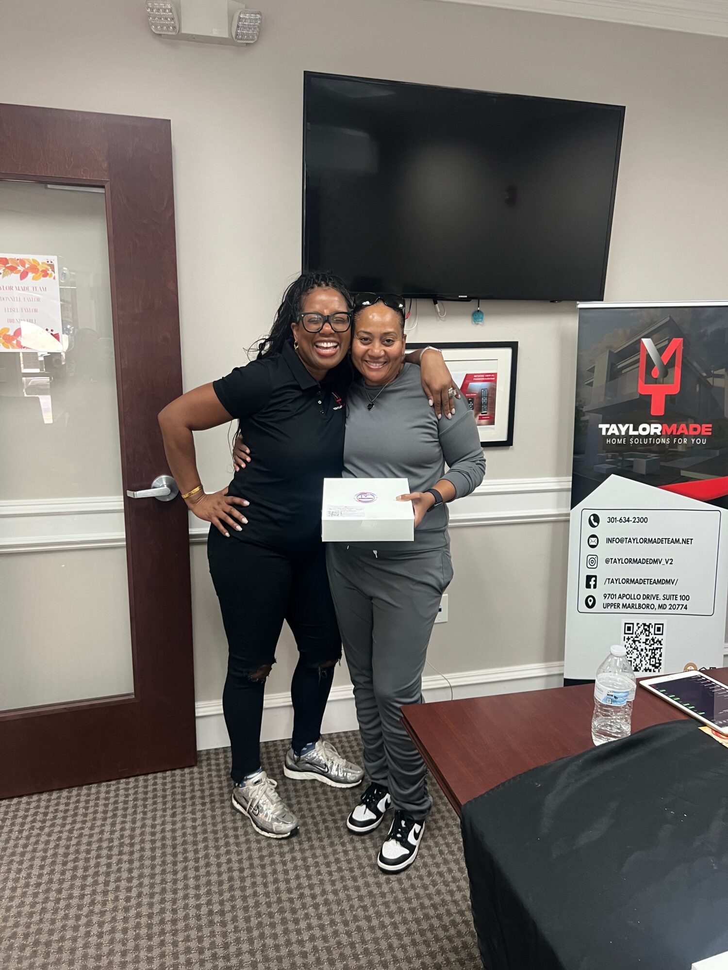 Two women standing together indoors, smiling, one holding a certificate, with a TV and TaylorMade display in the background.