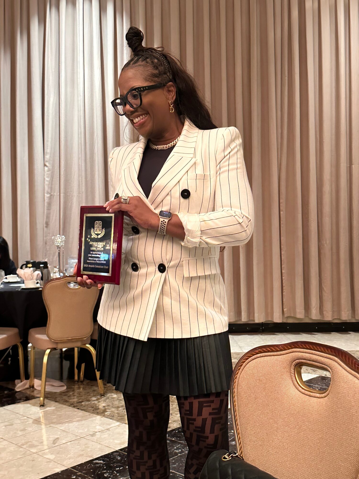 Woman in a pinstripe blazer holding a plaque, smiling, with curtains in the background.