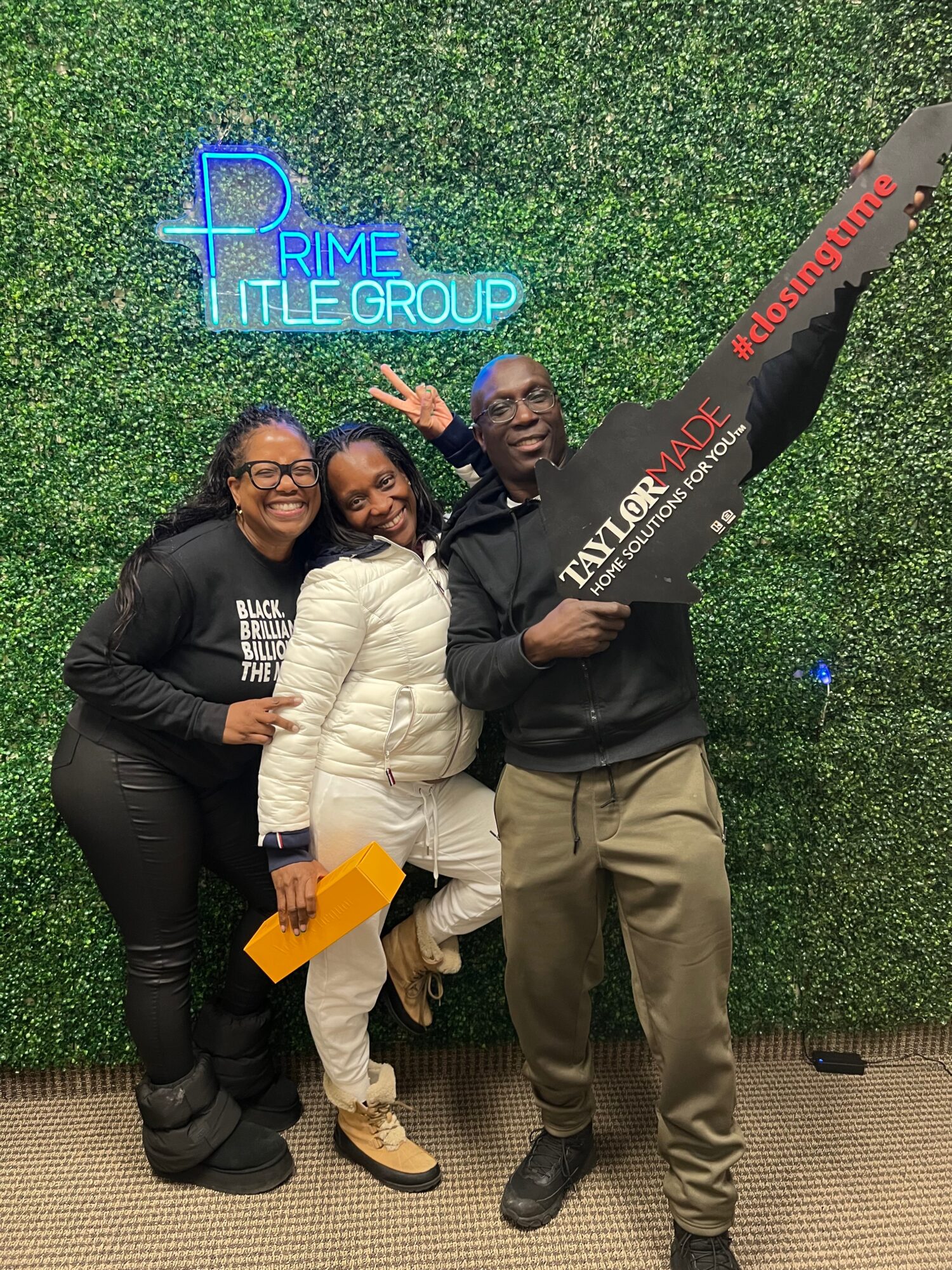 Three people smiling and posing in front of a green wall with a neon sign that reads 'Prime Title Group.'