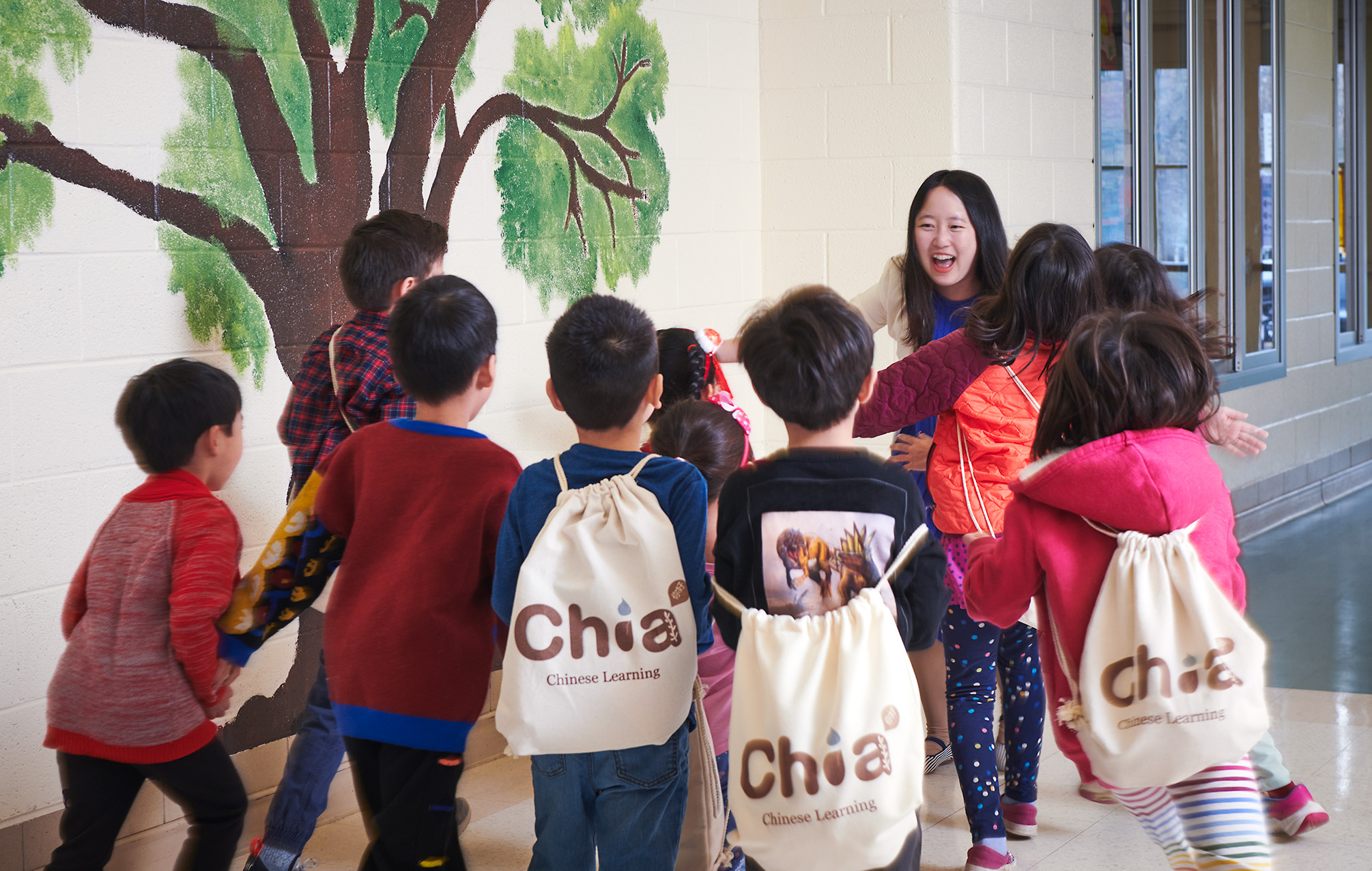 Group of children gathered around a woman near a painted tree mural, some carrying backpacks, indoors.