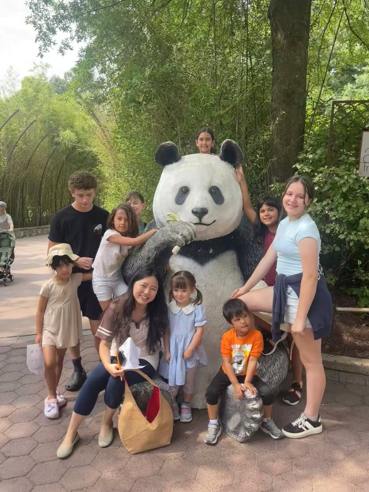 Group of children and adults with a person in a panda costume outdoors among trees, smiling and posing for photo.