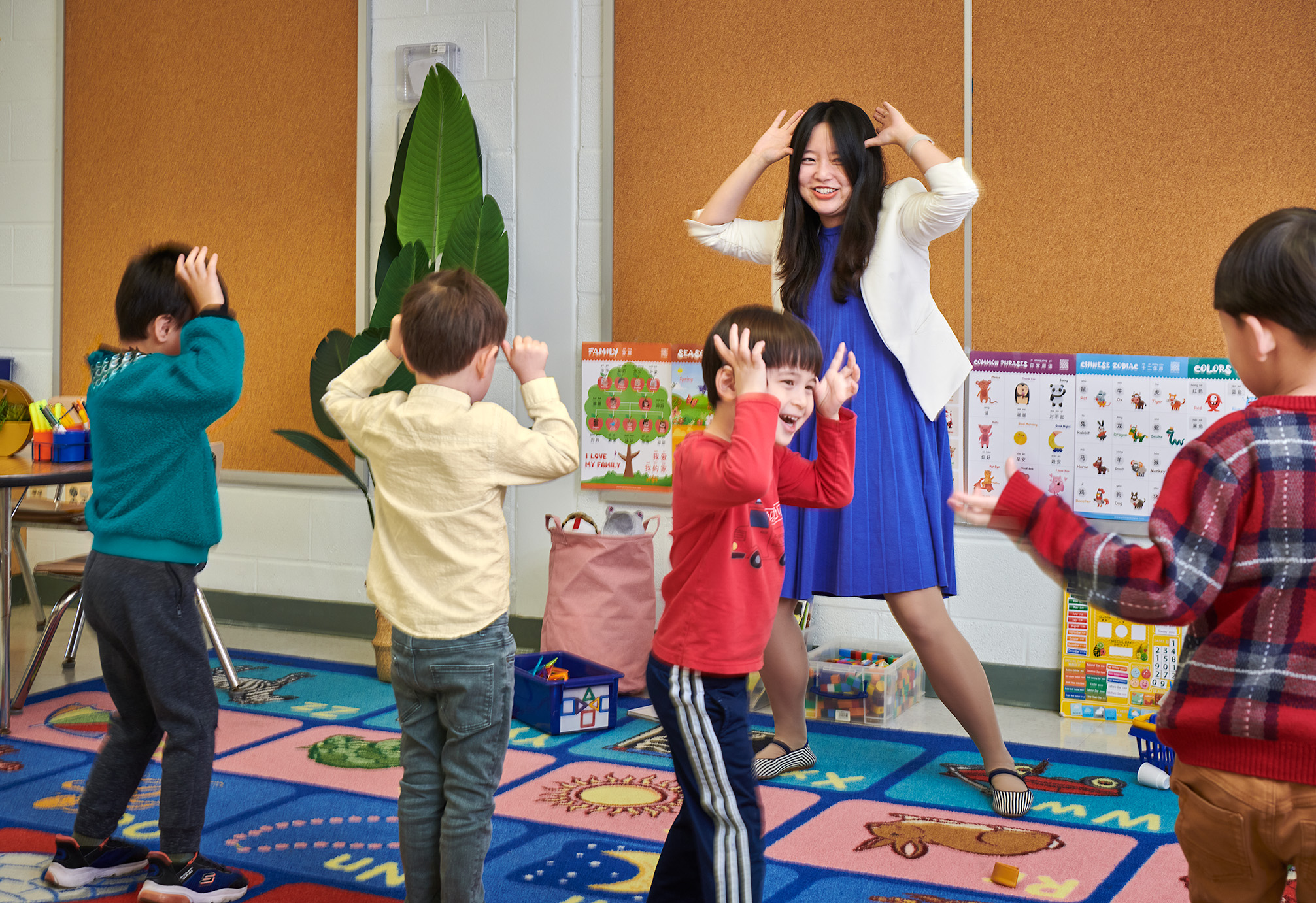 Teacher and children in a classroom, some with hands on heads, engaging in an activity, colorful educational posters on wall.
