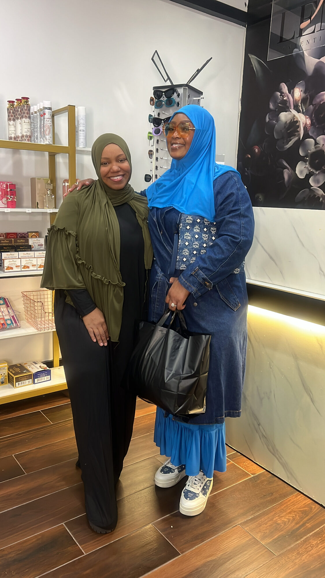 Two women standing indoors, smiling, one wearing a green jacket and black pants, the other in a blue hijab and dress, holding a black bag.