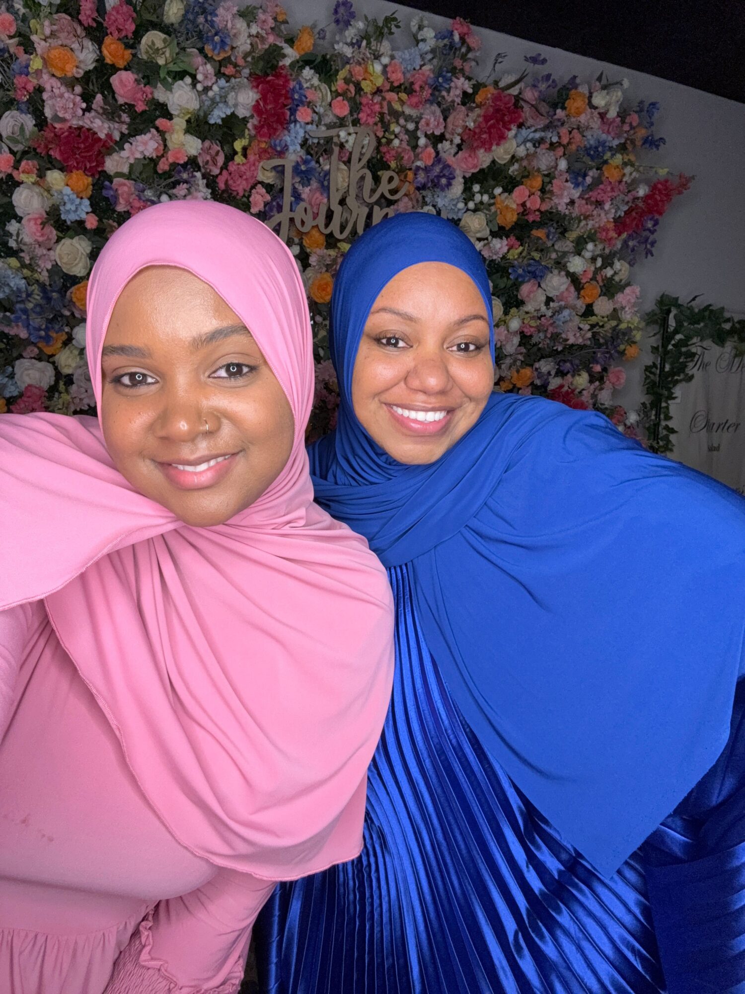 Two women wearing hijabs, one in blue and one in pink, smiling in front of a colorful floral background.
