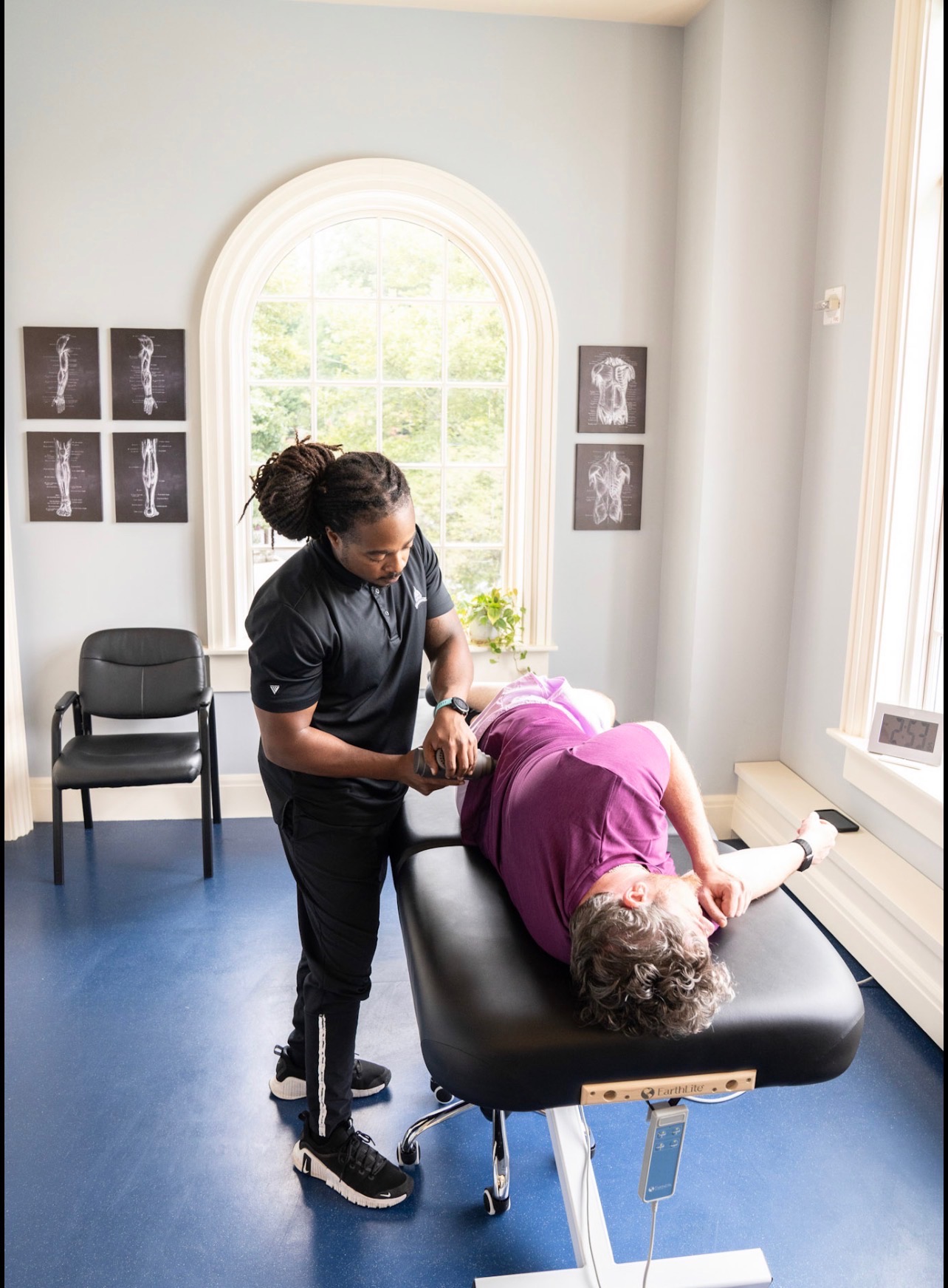 A healthcare professional performs a procedure on a patient lying on a treatment table in a bright room with a large window and framed images on the wall.