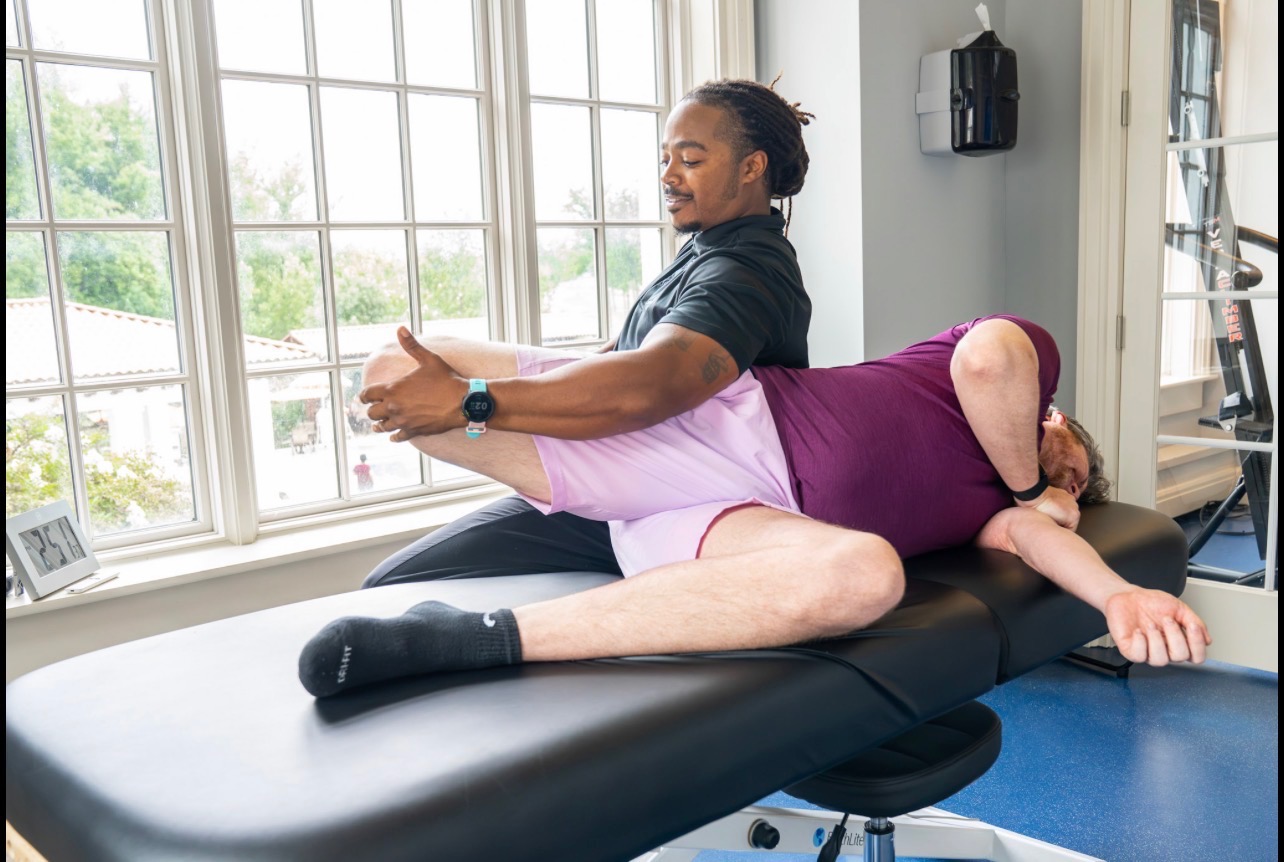 Therapist assisting a person with leg exercises on a treatment table in a bright room with large windows.